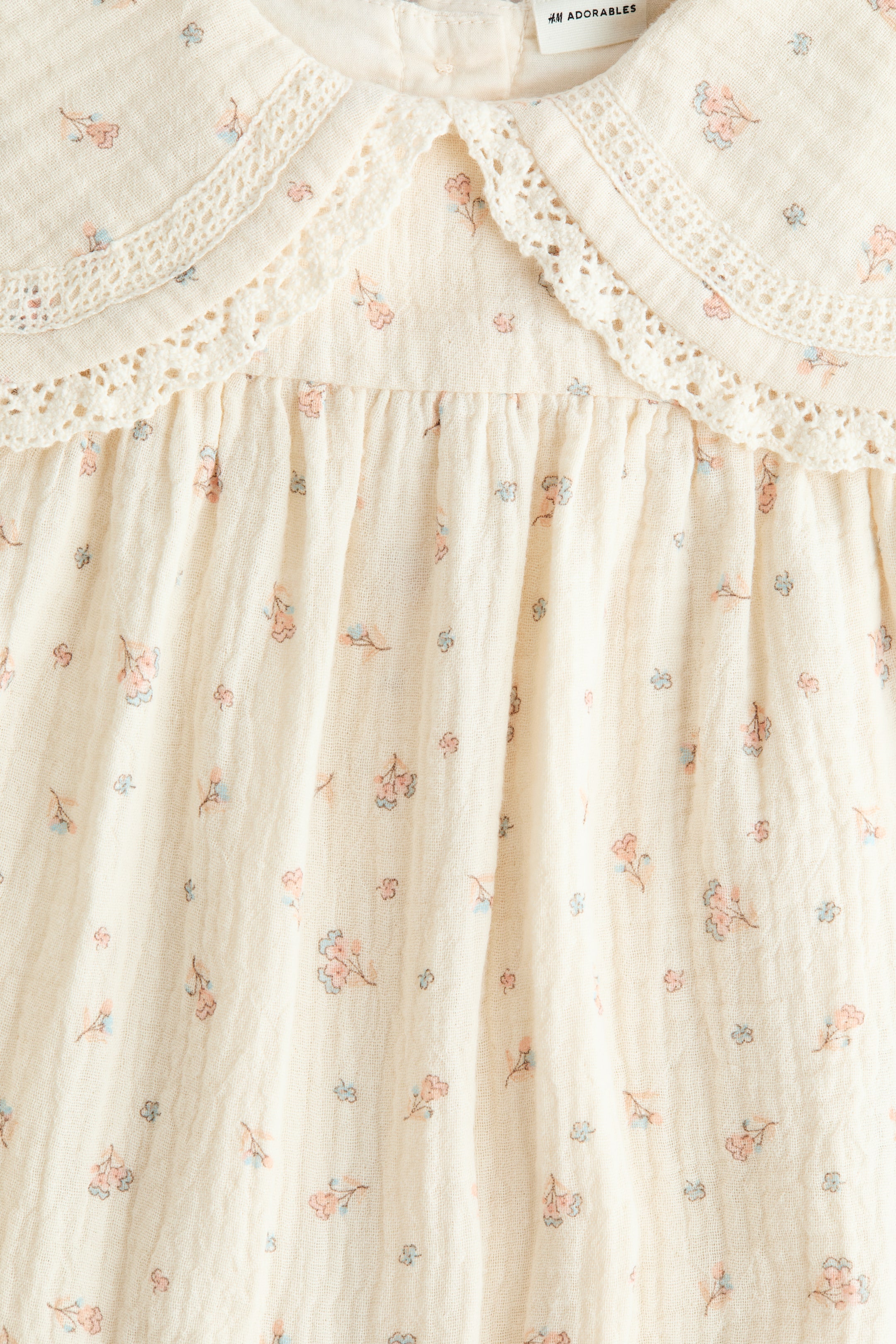 Ampliar la imagen: Una prenda de color crema está mostrando su frente, con un cuello Peter Pan con un ribete de encaje blanco. La tela, que presenta un pequeño patrón floral en rosa y azul, está visiblemente fruncida debajo del cuello y tiene una textura arrugada.