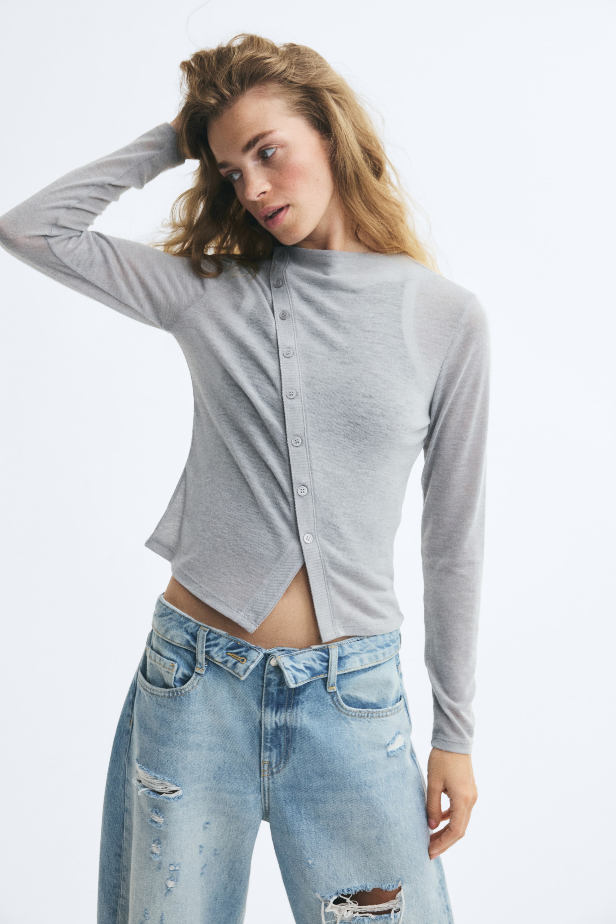 View larger image: A light grey, semi-sheer, fine-knit top is worn by the model, featuring a high neckline, long sleeves, and a diagonal ribbed button placket running down the front. The top is styled with light-wash distressed blue jeans.