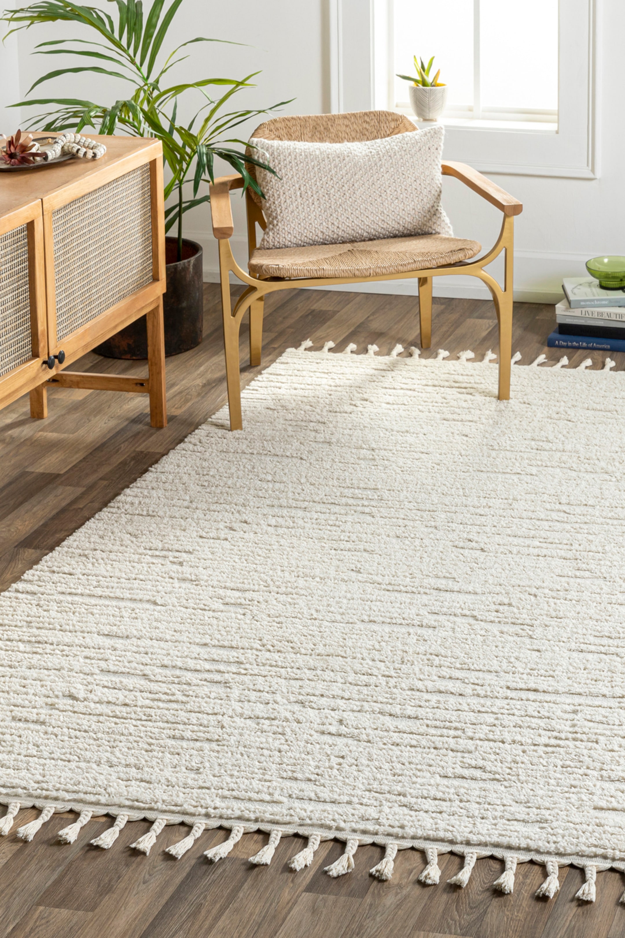View larger image: The cream-coloured rug features a textured surface with horizontal variations in pile height and is adorned with tassels along its visible edges. It lies flat, extending from the foreground towards the midground.