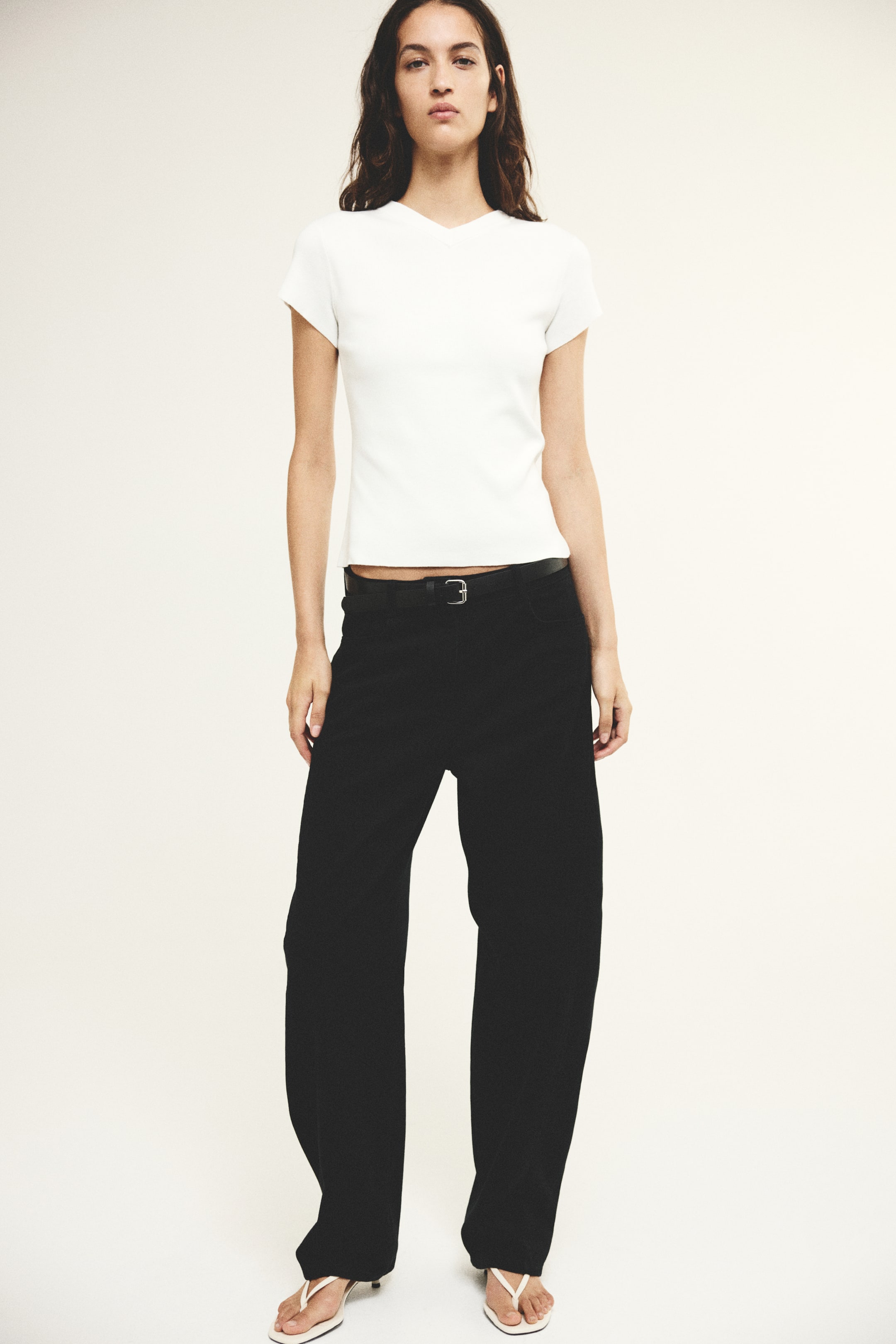 Grotere afbeelding bekijken: Zwarte high-waisted denim jeans met een gebogen, volumineuze pijp en een bijpassende zwarte riem met een zilverkleurige gesp worden gedragen door het model. De jeans zijn gestyled met een aansluitend wit T-shirt met korte mouwen en witte sandalen met hak en teenslippermodel.