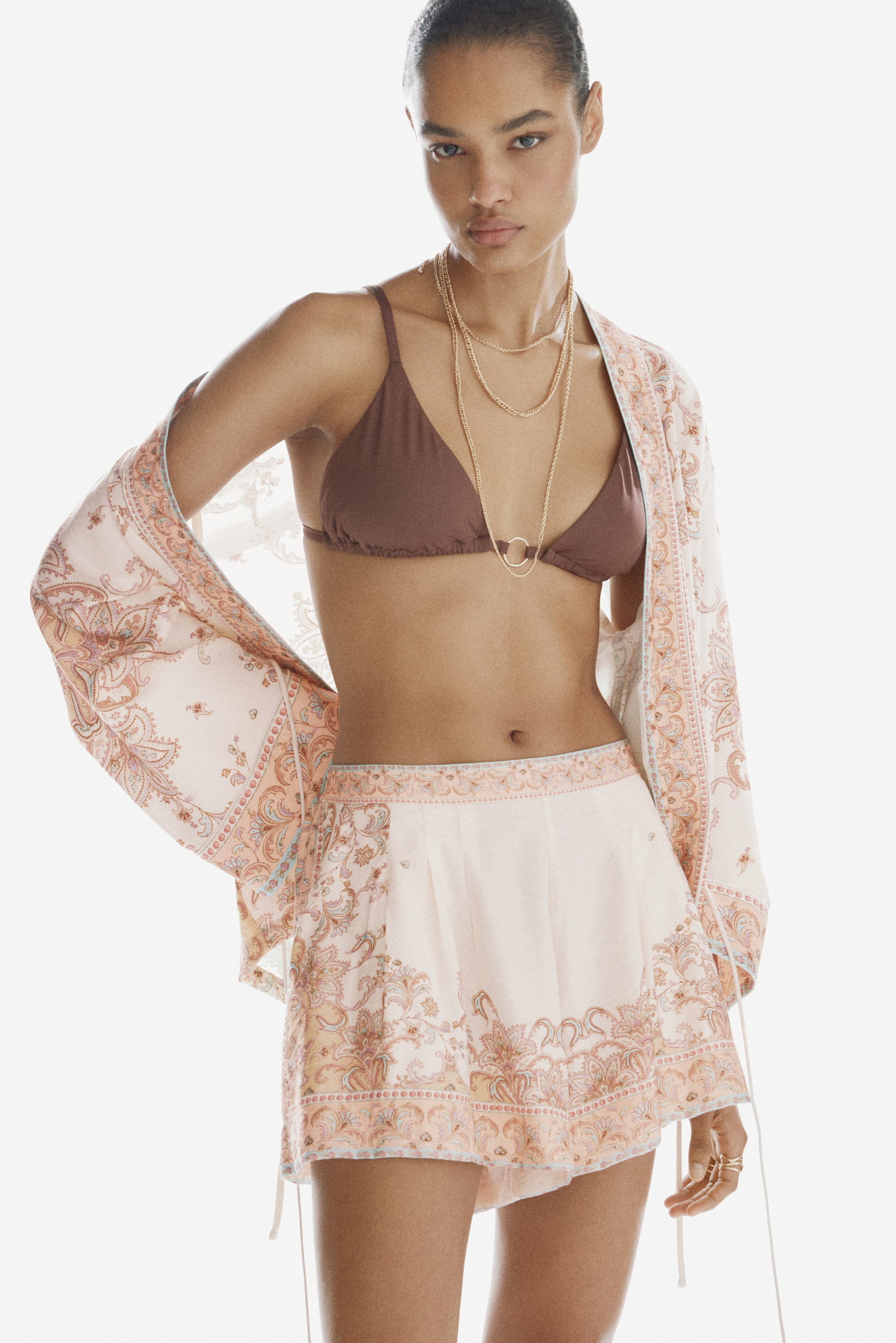 View larger image: The model is wearing loose-fit shorts with an all-over paisley print and narrow piping. These shorts have a regular waist with pleats at the top and diagonal side pockets. The model is also styled in a matching sheer kimono.