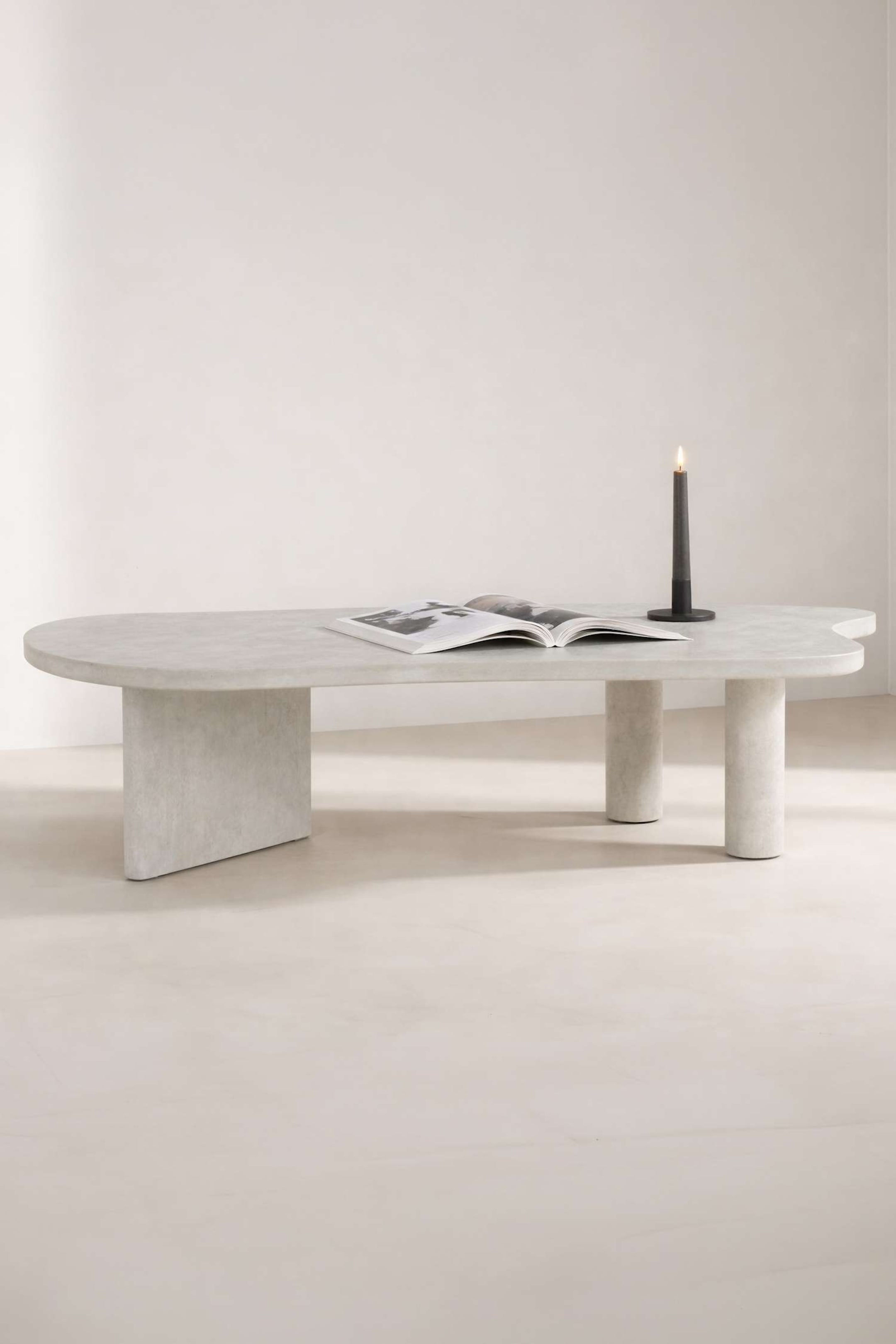 Agrandir l'image: Une table basse gris clair présente un plateau ovale allongé et irrégulier, légèrement orienté vers la droite. Elle est soutenue par trois pieds distincts : l’un est une dalle rectangulaire plate, et les deux autres sont des piliers cylindriques. Un livre ouvert et une bougie allumée reposent sur sa surface.
