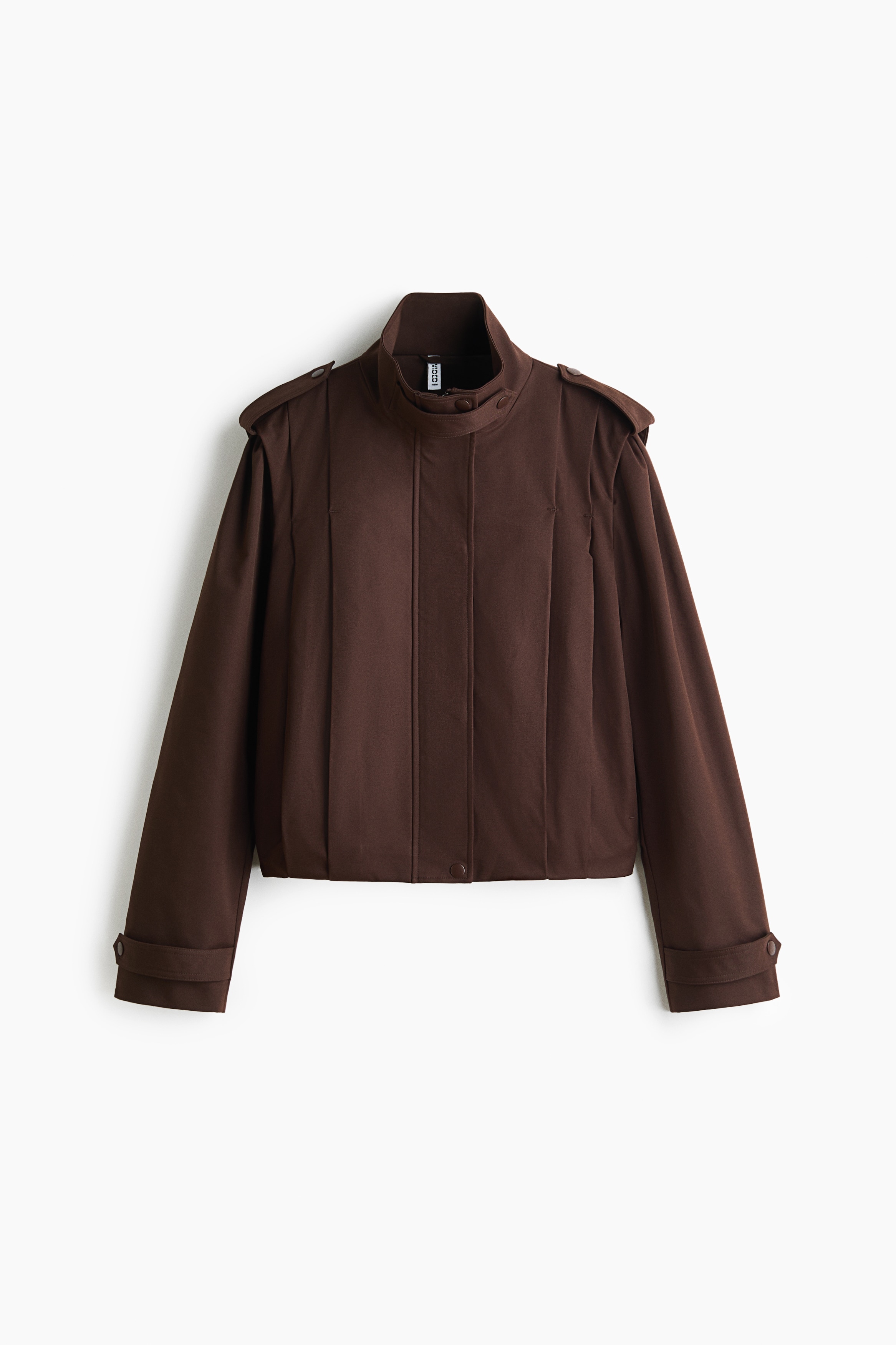 A short, oversized dark brown twill jacket featuring a high funnel collar with a throat latch, epaulets, and a pleated back is being worn by the model. The model is sitting while wearing the jacket paired with a patterned skirt.