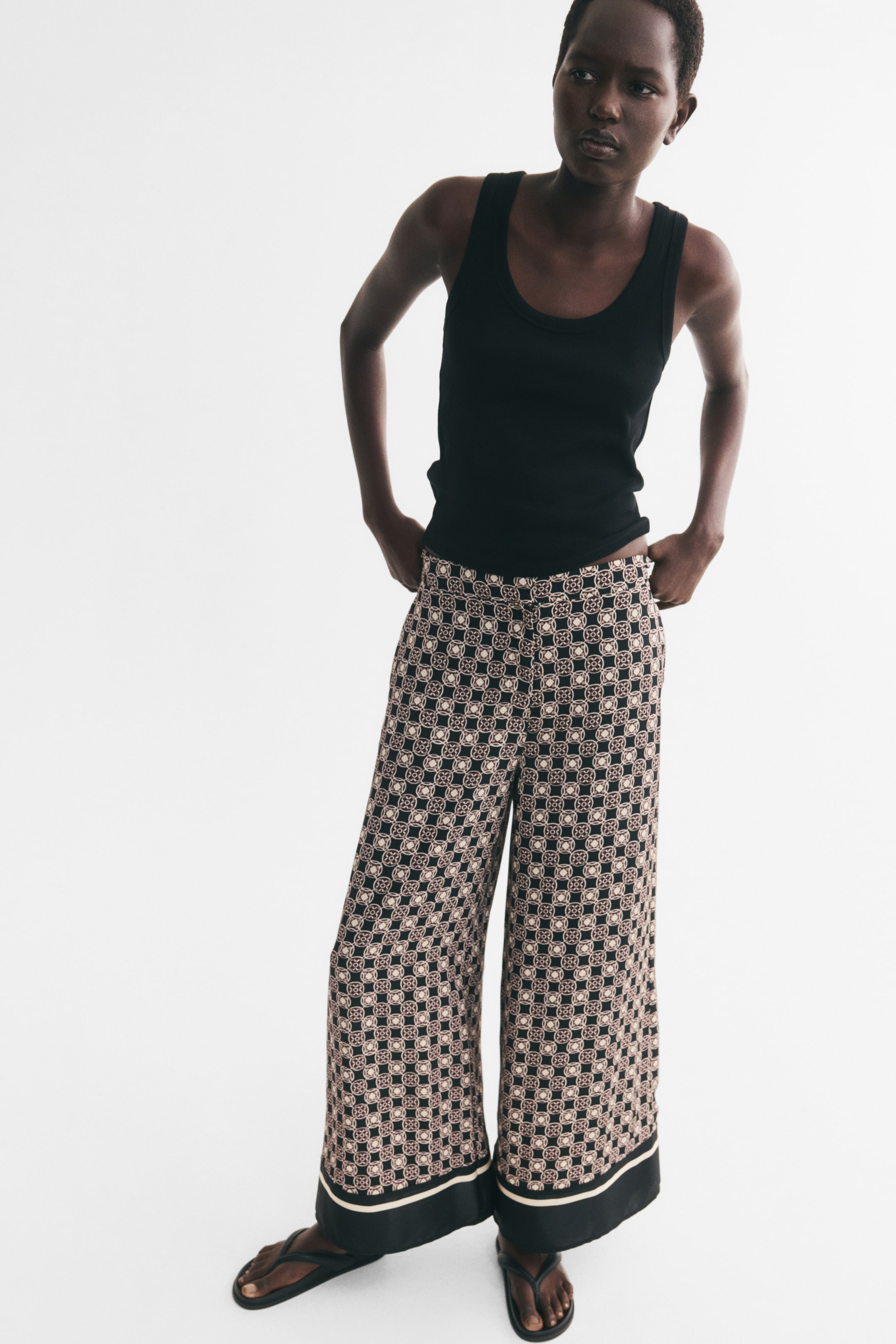 View larger image: Wide-leg pants with a geometric pattern and a solid black hem accented by a cream stripe are worn by the model, who is resting their hands on the waistband. The high-waisted pants are styled with a black tank top and black thong sandals.