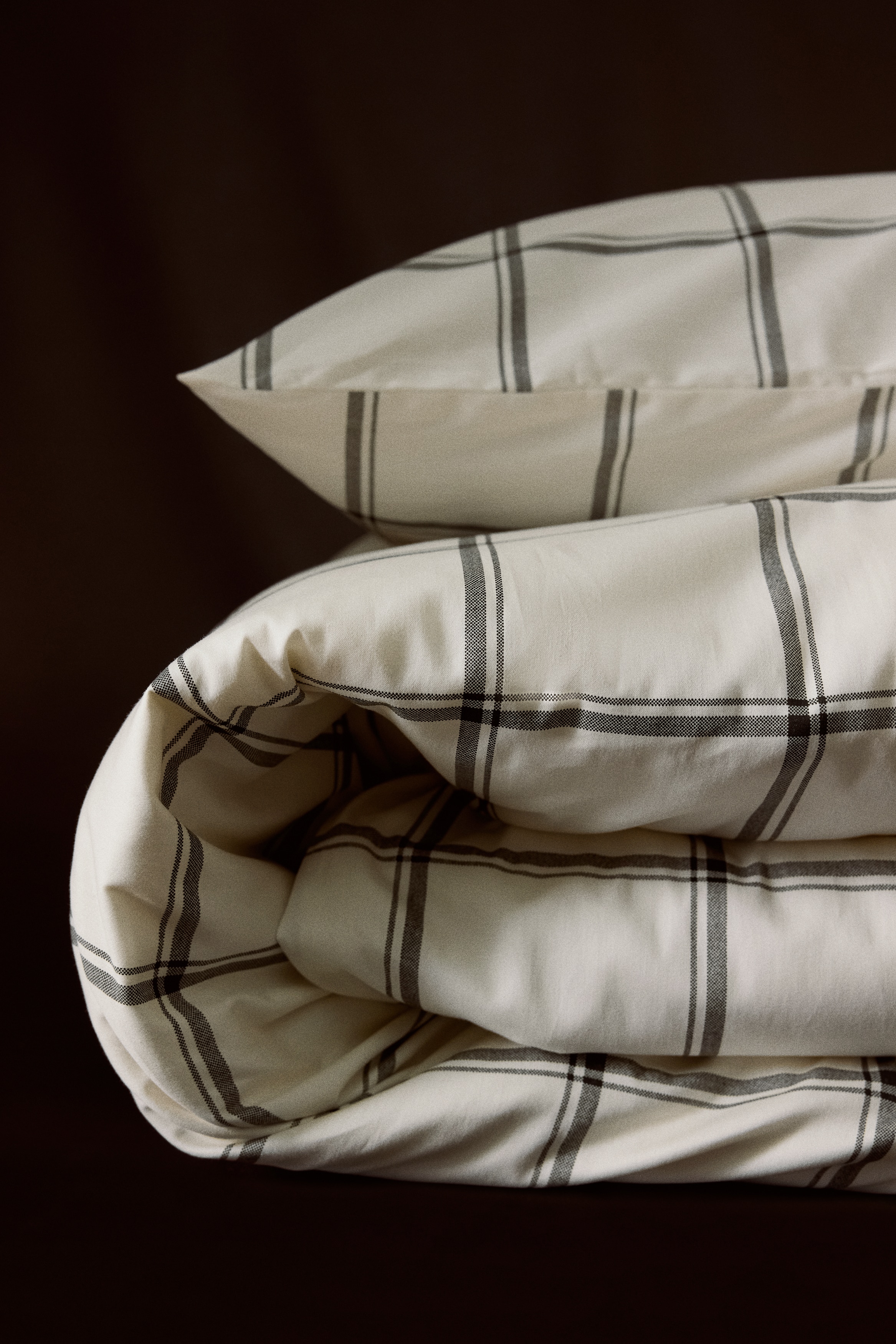 Un ensemble de housse de couette présente une couleur crème avec un motif à carreaux noirs, comprenant une taie d'oreiller debout derrière une housse de couette pliée. Les deux articles montrent leurs faces avant, affichant le motif à carreaux dans son intégralité.