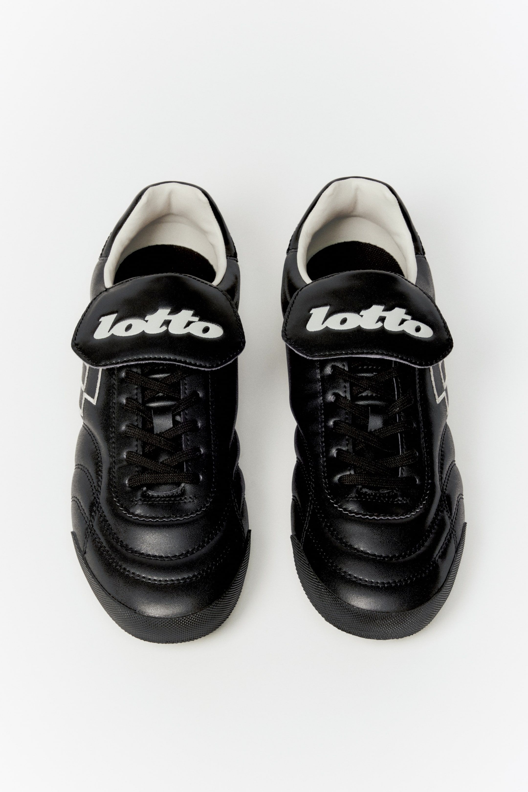 View larger image: A pair of black athletic shoes sits side-by-side, facing forward, showcasing black laces and large tongue flaps with "lotto" in white script. These shoes feature quilted stitching across the toe boxes, white linear detailing on the sides, and a white lining around the collar.