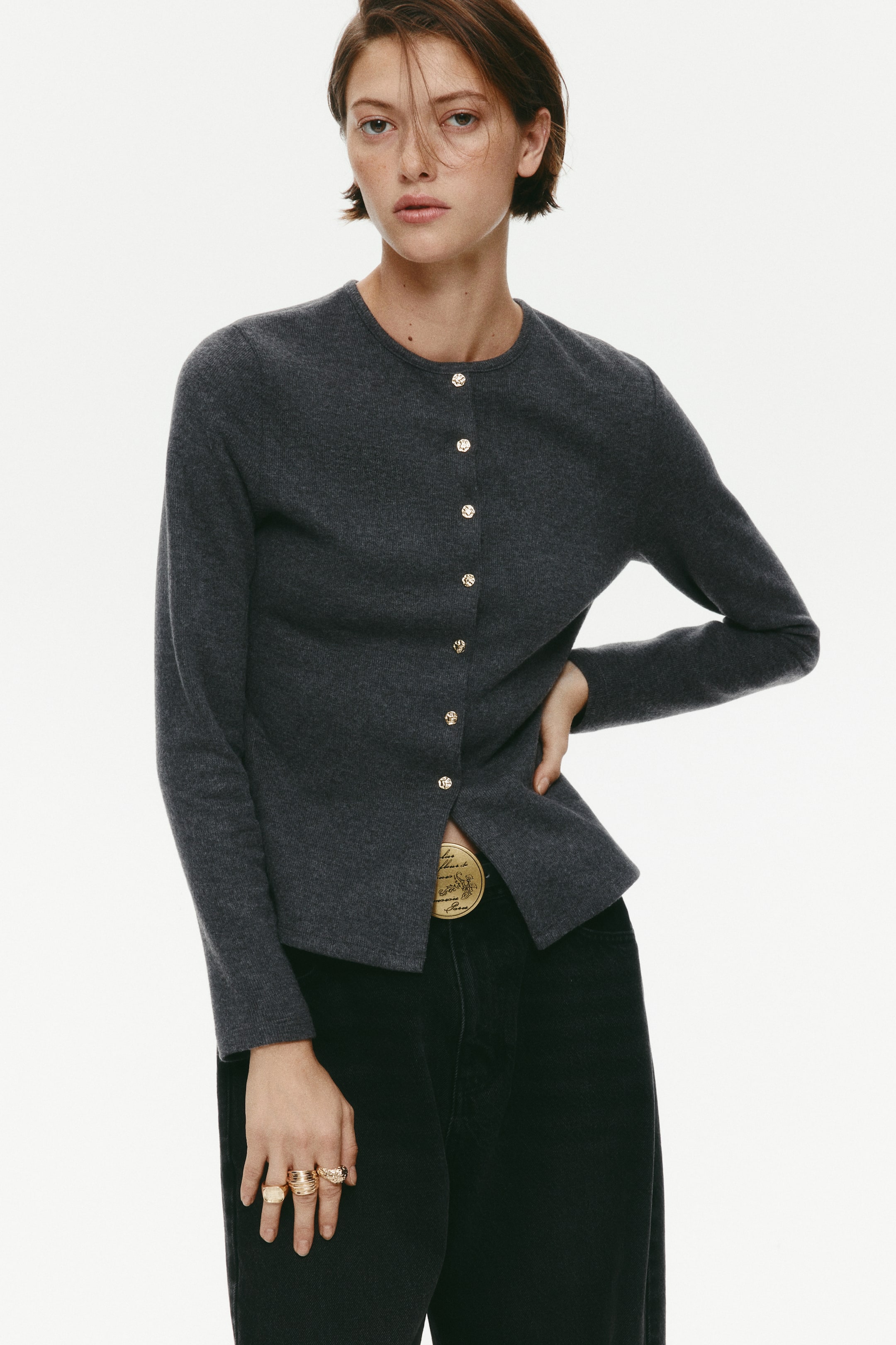 View larger image: A dark grey cardigan in a soft, fine knit is being worn by the model, featuring a round neckline, long sleeves, and irregular gold-toned metal press studs down the front. The cardigan is styled with the lower studs left unfastened to show black trousers and a gold belt buckle.