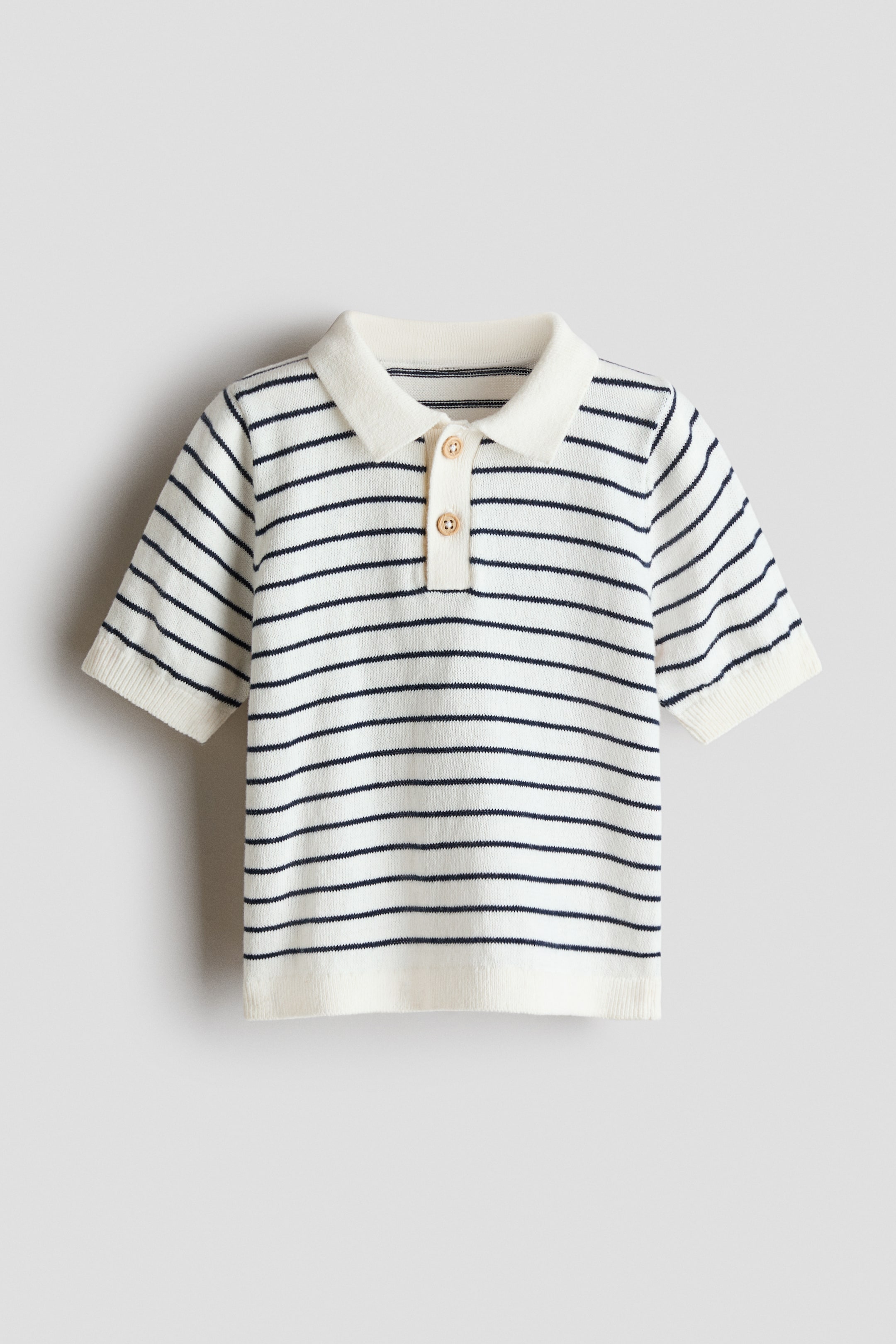 Agrandir l'image: Un polo en tricot est présenté de face, affichant des rayures horizontales blanc cassé et bleu marine. Il est doté d'un col, d'une patte de boutonnage à deux boutons avec des boutons en bois clair, de manches courtes, ainsi que de poignets et d'un ourlet côtelés.