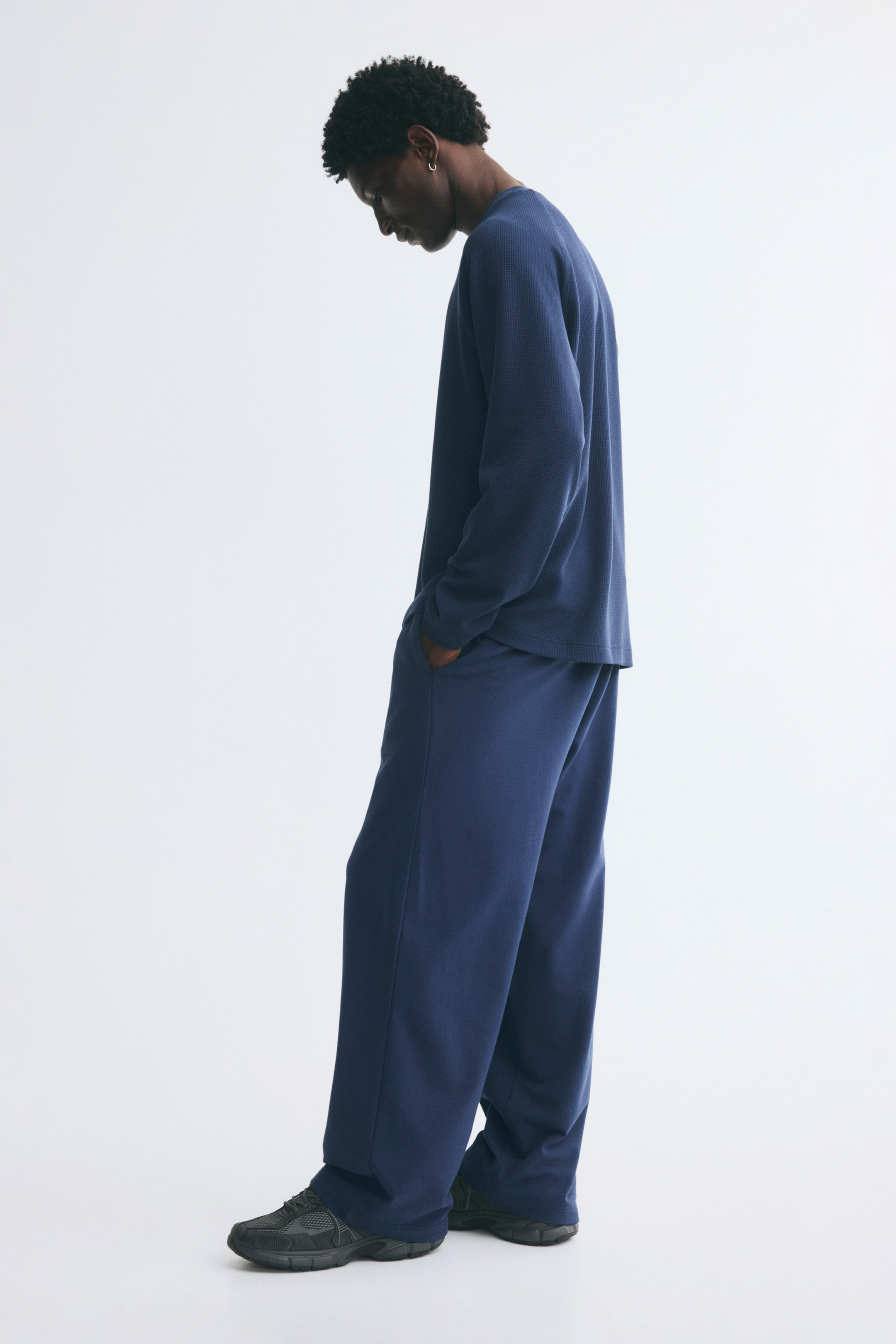 Voir une image plus grande: Un pantalon de sport bleu à coupe ample et jambes larges est porté par le modèle, qui se tient de profil avec une main glissée dans une poche latérale. Le pantalon est agencé avec un haut bleu à manches longues assorti et des sneakers foncées.