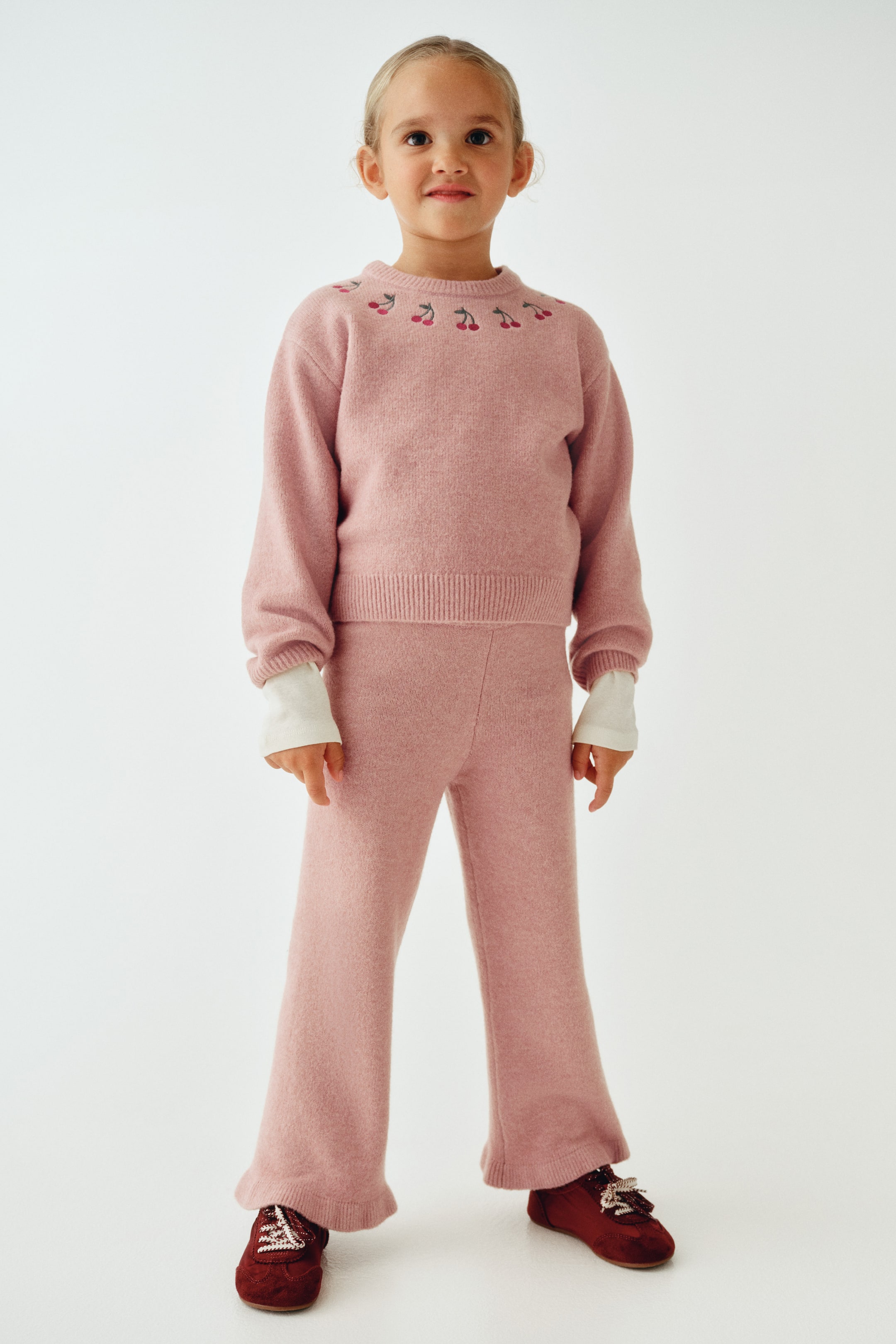 Visualizza l'immagine ingrandita: Un bambino indossa un completo in maglia rosa polveroso, mostrato frontalmente, composto da un maglione a maniche lunghe con dettagli di ciliegie ricamate attorno allo scollo a coste e pantaloni in maglia coordinati a gamba dritta. Il maglione è sovrapposto a un top bianco con polsini in vista, e i pantaloni abbinati presentano un orlo a coste.