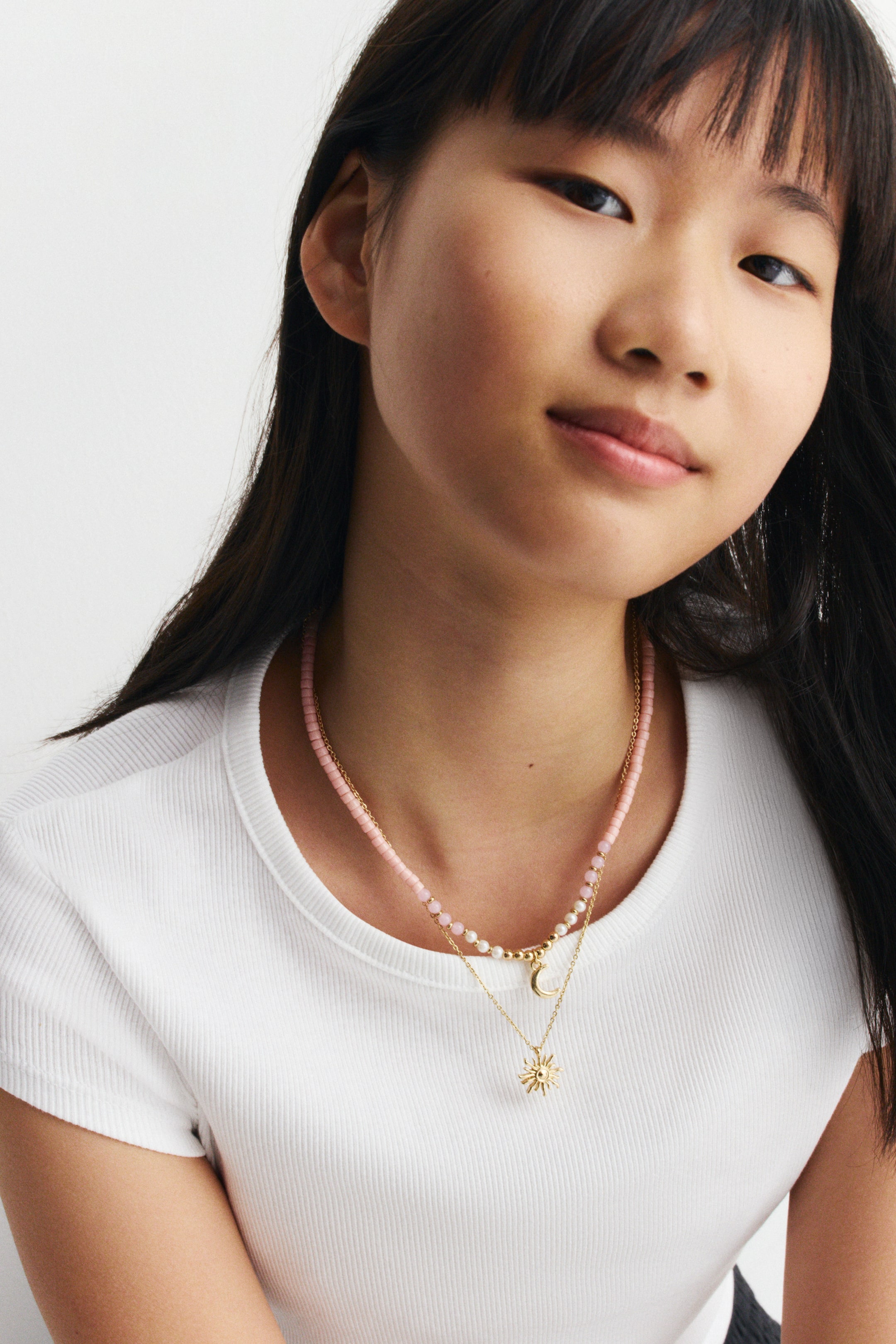 Ver imagen más grande: Un conjunto de collares en capas se lleva puesto, con una cadena corta de pequeñas cuentas rosas. Una segunda cadena de tono dorado se superpone debajo, adornada con cuentas blancas y un dije de luna creciente, mientras que una tercera cadena más larga de tono dorado cuelga más abajo con un dije de destello solar. Los collares descansan de frente sobre una camiseta blanca de canalé.