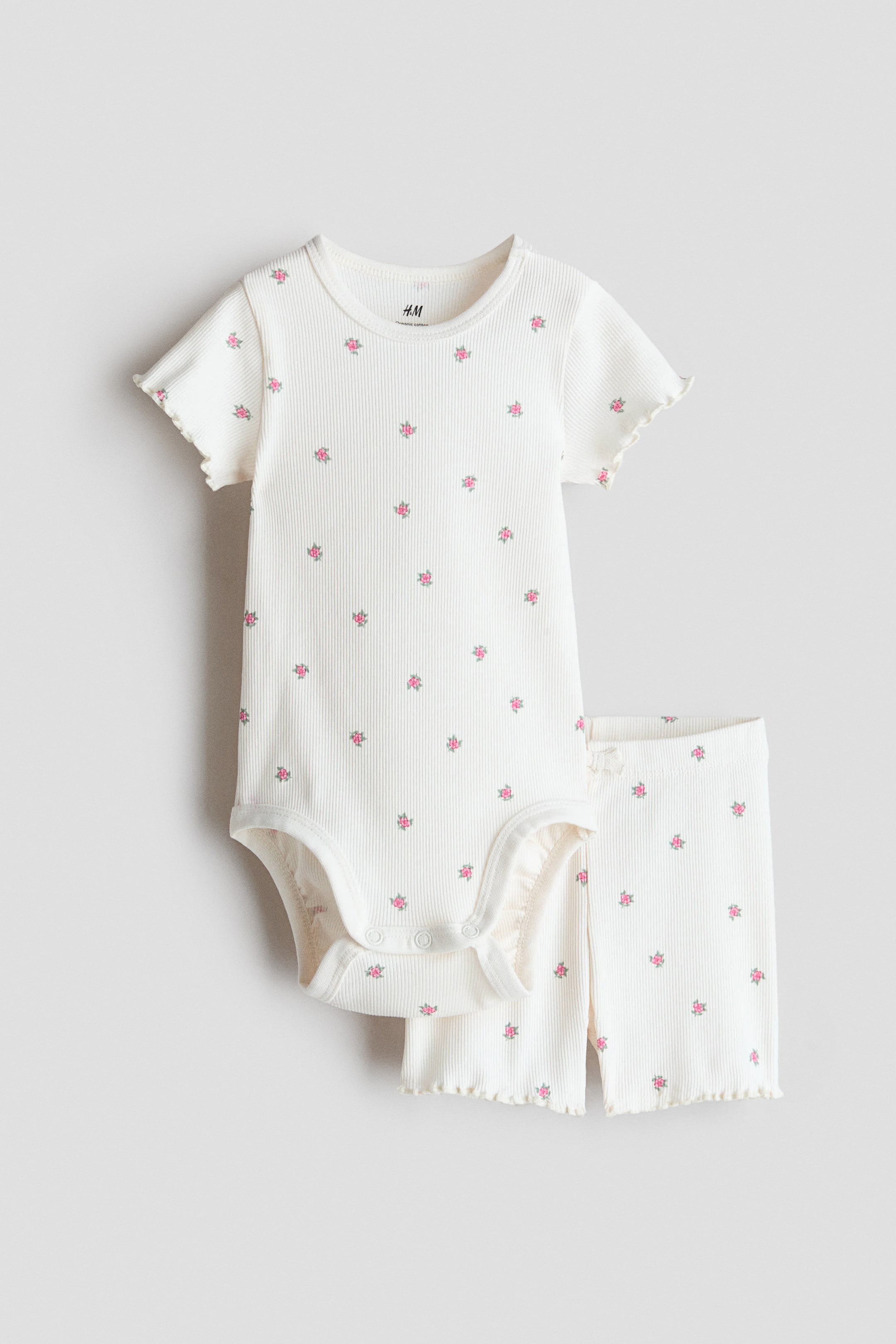 View larger image: An off-white, rib-knit baby onesie and matching long pants set features a pattern of small pink and gray floral motifs. The onesie has short sleeves with ruffled edges and snap closures at the crotch, while the coordinating pants display a ruffled hem.