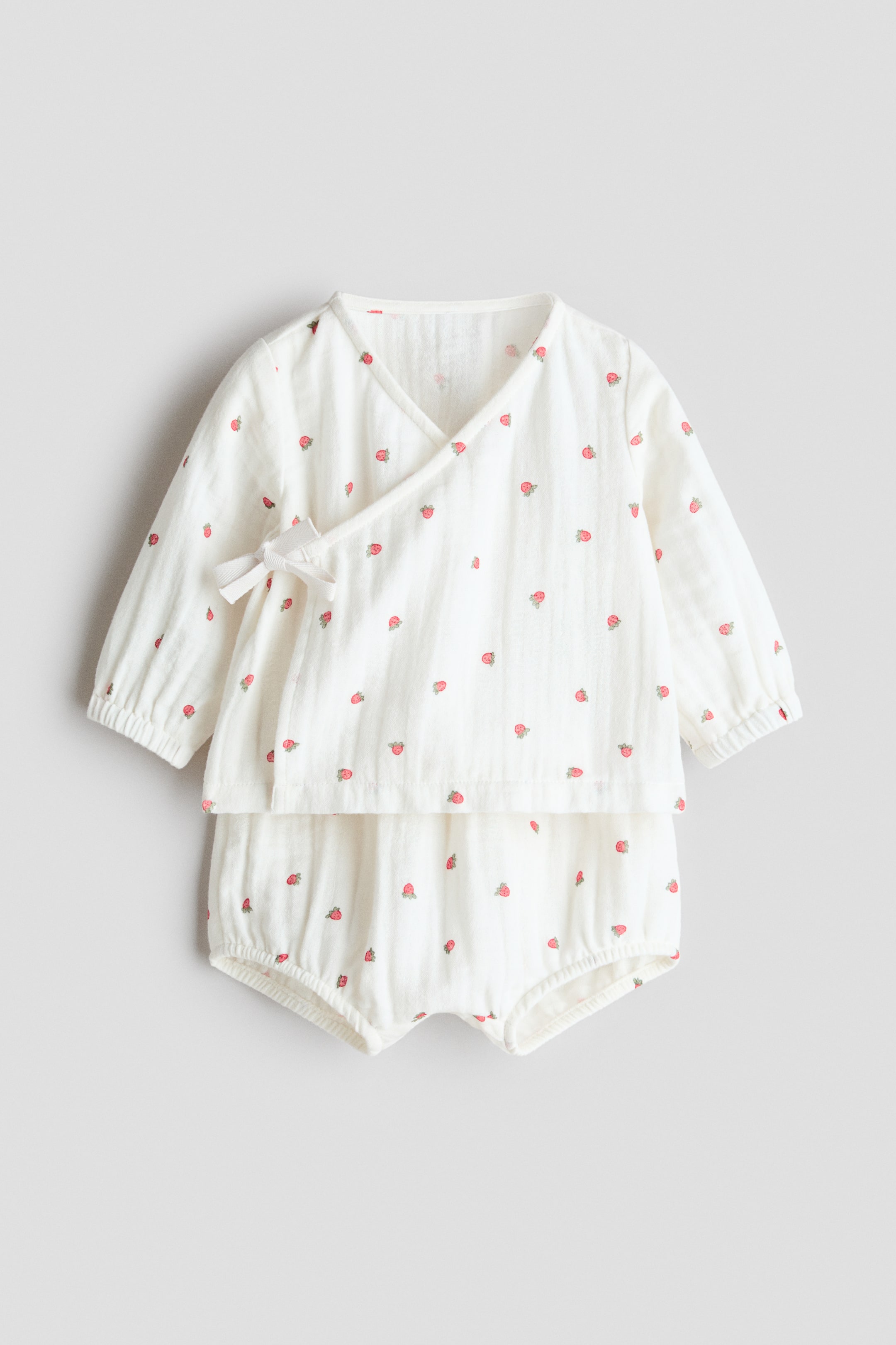 View larger image: A cream-coloured baby clothing set is presented at the front, comprising a top and bloomers in a textured fabric adorned with an all-over strawberry print. The top features a V-neck, long sleeves with elasticated cuffs, and a wrapover front with a decorative bow tie. The bloomers have elasticated leg openings.