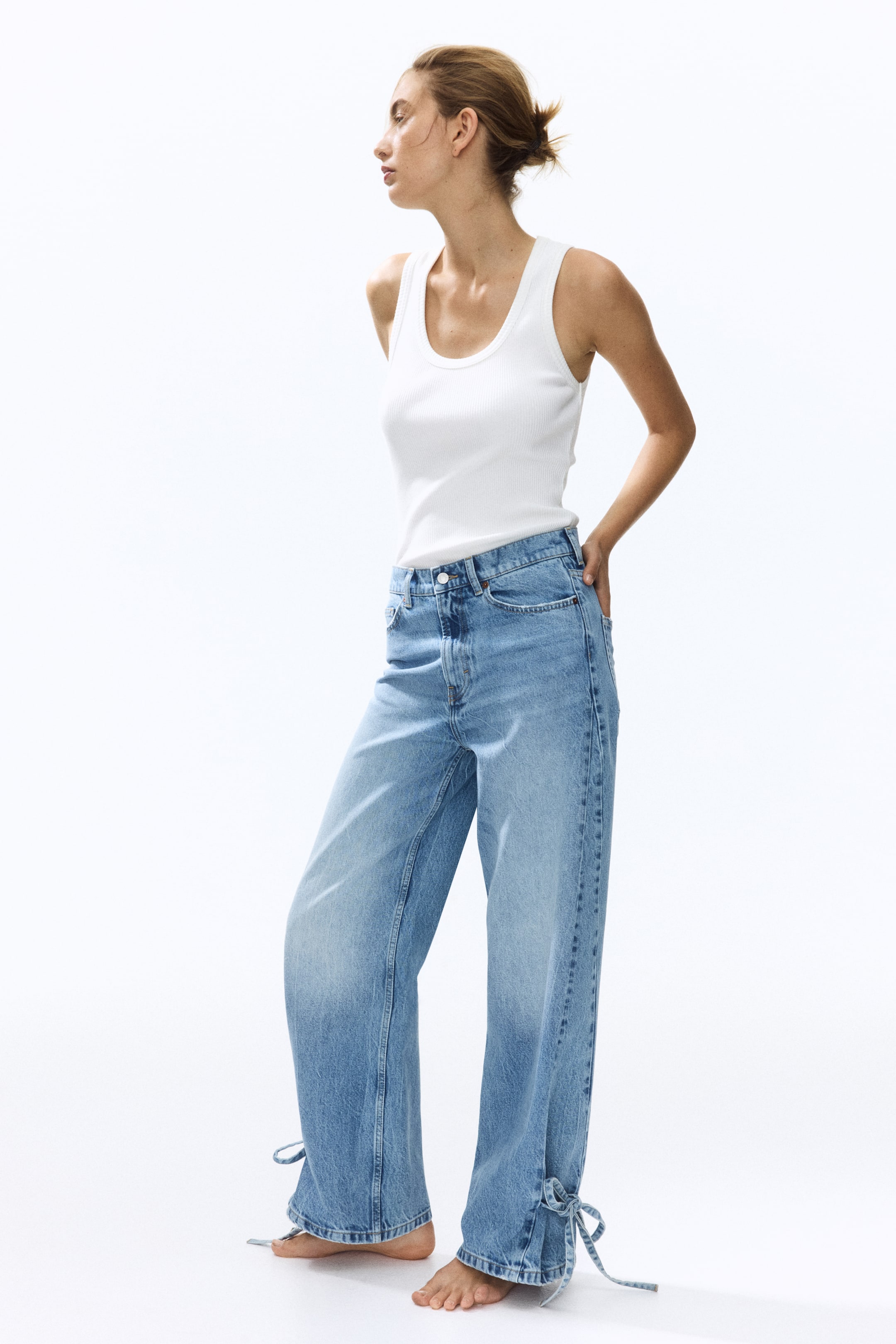 Visa större bild: Ljusblåa 5-ficksjeans med hög midja, dragkedjegylf och knappstängning bärs av modellen, som också har på sig ett vitt linne. Jeansen har böjda, voluminösa ben som avsmalnar mot de ankel-långa fållarna, där smala band är synliga. Modellen placerar en hand i en bakficka.