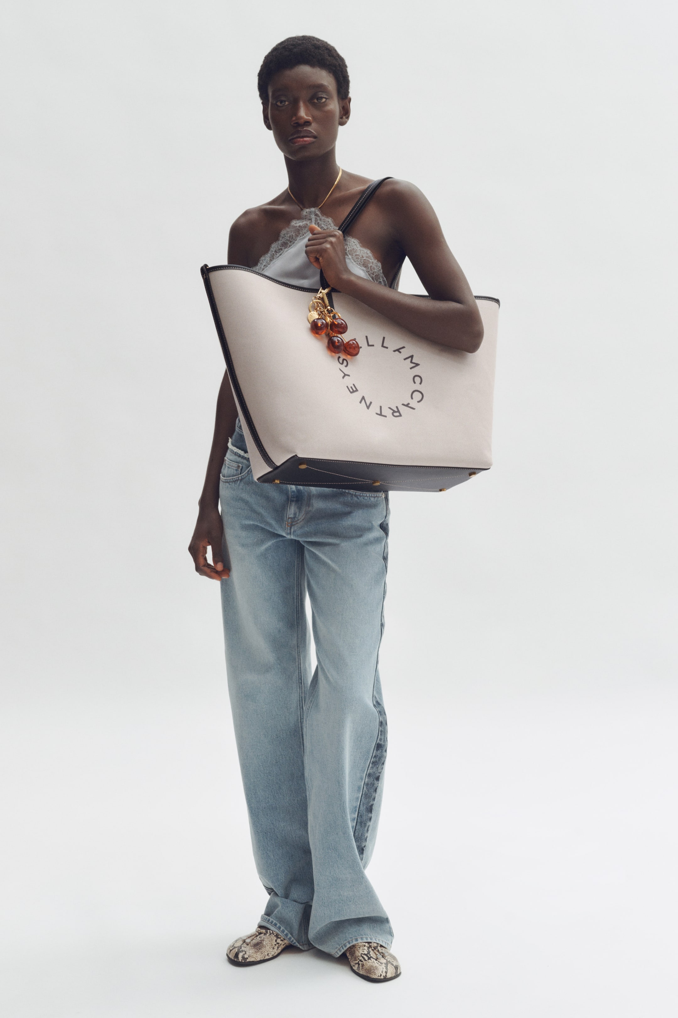 Agrandir l'image: Un sac fourre-tout en toile de couleur crème est tenu à un léger angle, mettant en valeur son devant avec un logo circulaire noir 'Stella McCartney'. Il présente une garniture contrastante foncée le long de ses bords et de son fond, et des breloques en forme de cerises ambrées pendent de sa bandoulière foncée.