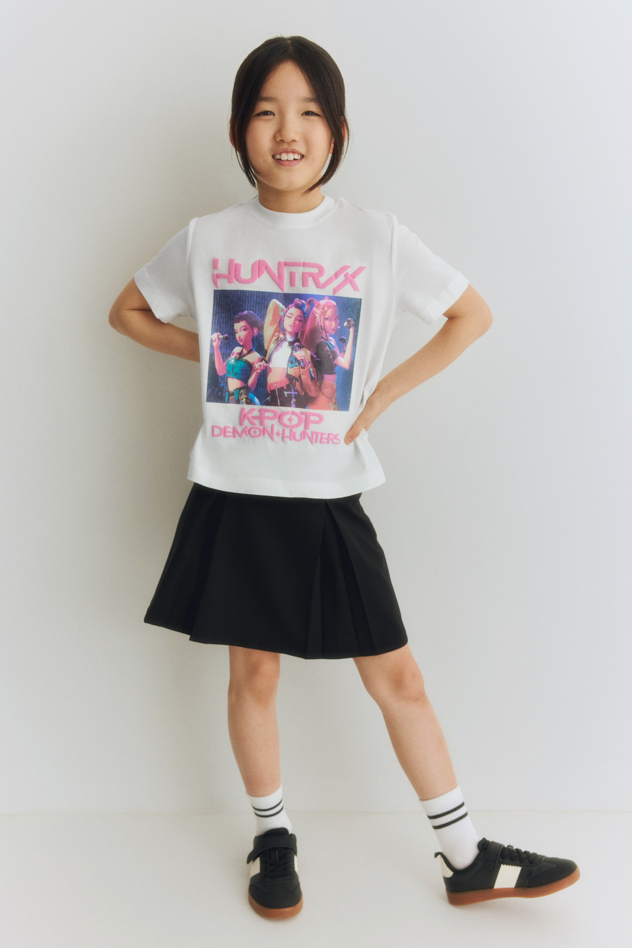 View larger image: The front of a white t-shirt showcases a graphic print of three female figures, with "HUNTRXX" in pink lettering above and "KPOP DEMON HUNTERS" in pink lettering below. It features a round neckline and short sleeves.