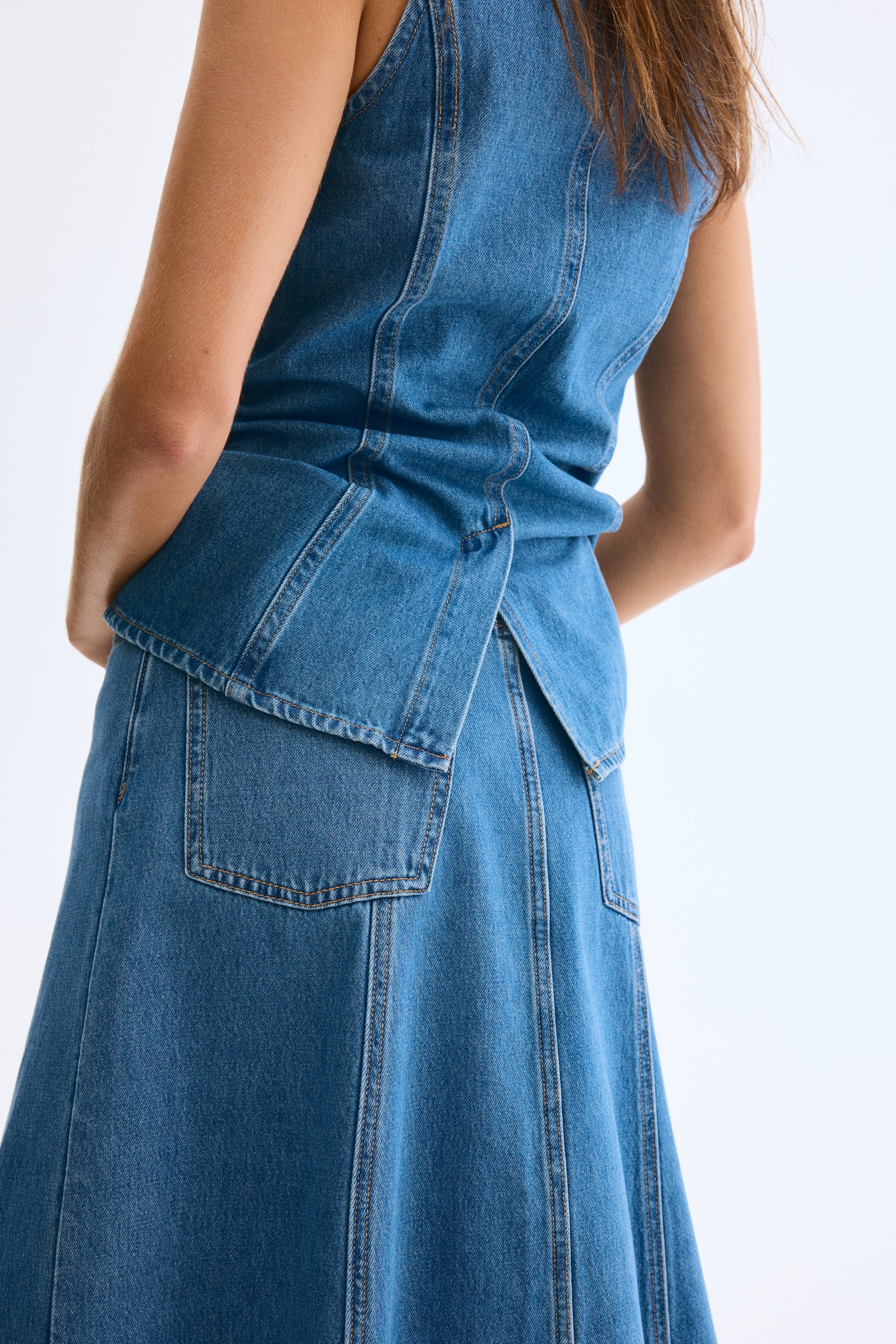 View larger image: A sleeveless denim top and A-line skirt are being presented as a coordinating set, viewed from the back. The top is featuring prominent vertical seams and a layered panel at the waist, which is creating a central back split, and the skirt is showcasing matching vertical seams and back patch pockets.