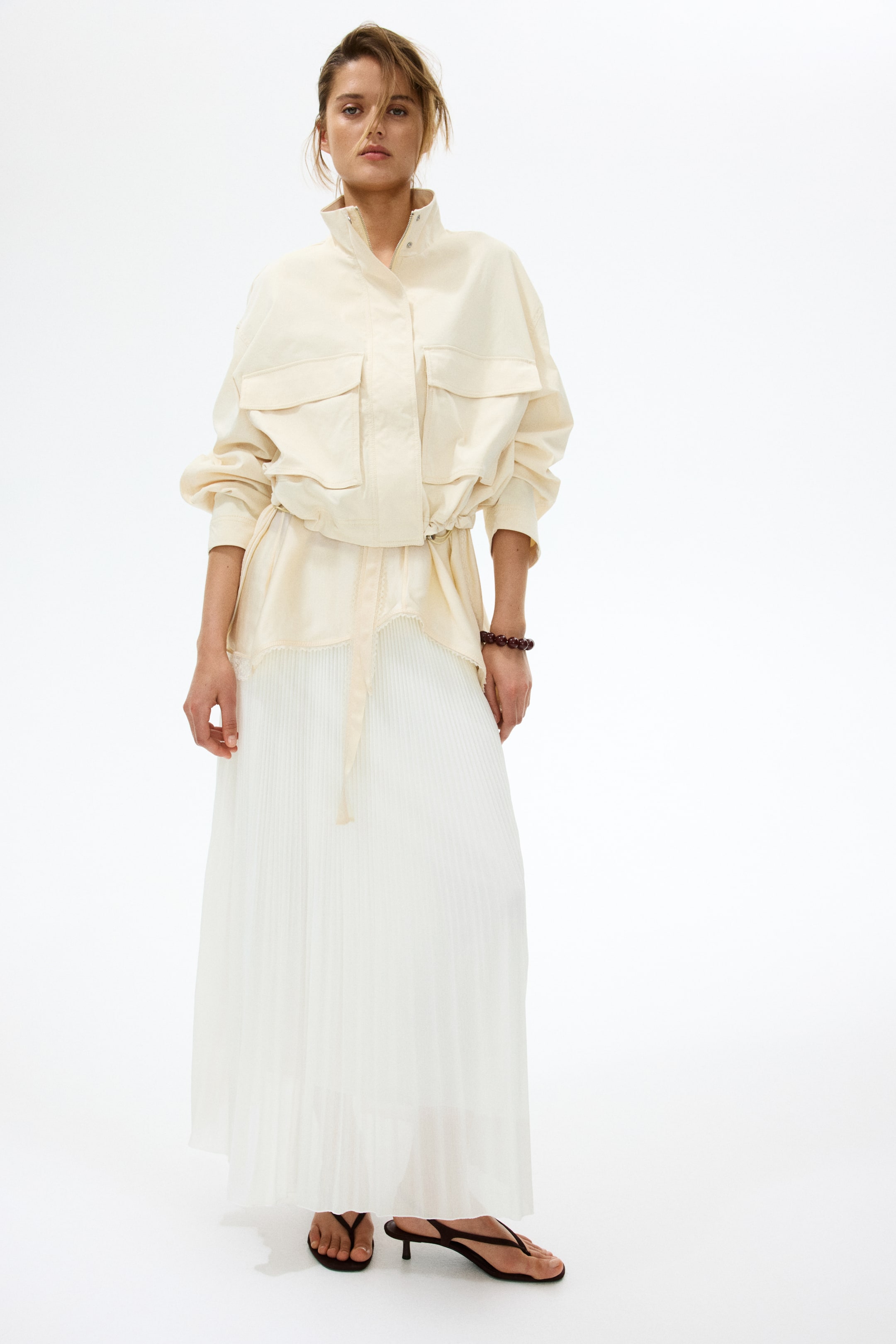 Ver imagen más grande: Una chaqueta utilitaria color crema presenta un cuello alto, bolsillos con solapa en el pecho y mangas voluminosas con puños fruncidos, su cintura con cordón se ciñe sobre una falda maxi plisada blanca. Se llevan sandalias de dedo marrón oscuro con tacón bajo, y una pulsera de cuentas adorna la muñeca derecha.