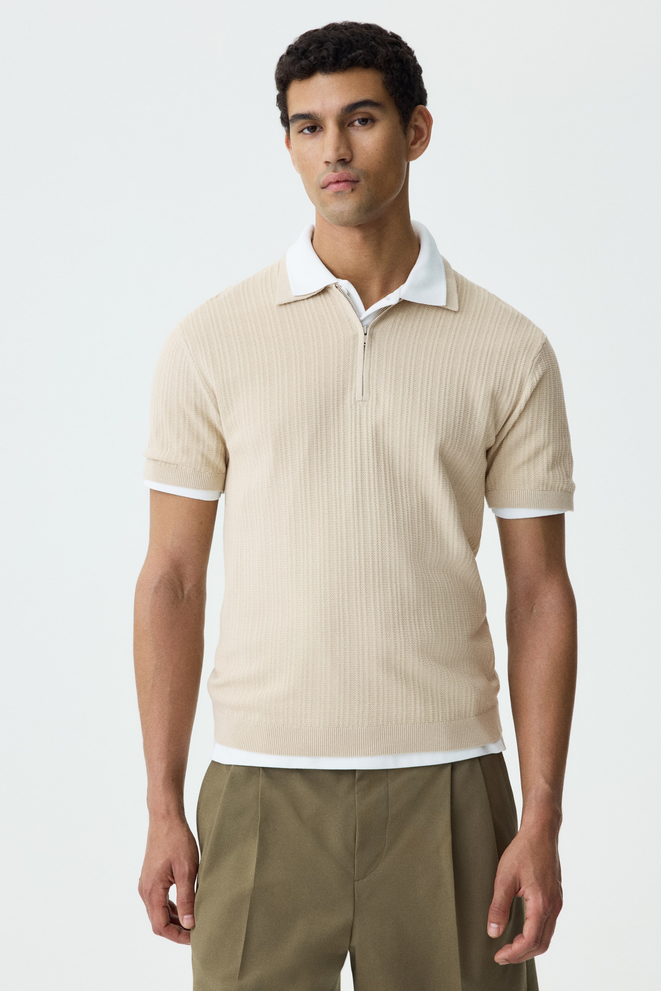 View larger image: The model is showcasing a beige textured knit polo shirt, accentuated by a white collar and a partial zip at the top. This slim-fit shirt boasts short sleeves with rib-trimmed cuffs and a rib-trimmed hem, subtly revealing a white undershirt beneath. Perfectly crafted for a refined look, this piece is ideal for those who appreciate understated elegance in their wardrobe.