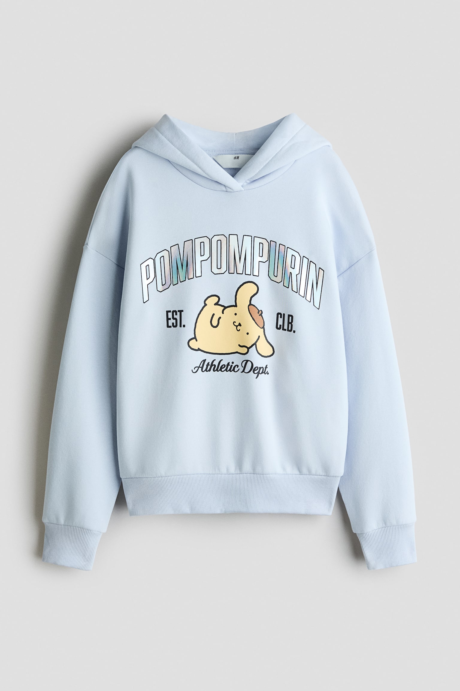 A light blue oversized hoodie is shown from the front, featuring a wrap hood, dropped shoulders, and ribbed cuffs and hem. The chest displays a printed graphic of the cartoon character Pompompurin beneath arched âPOMPOMPURINâ lettering and above the script âAthletic Dept.â