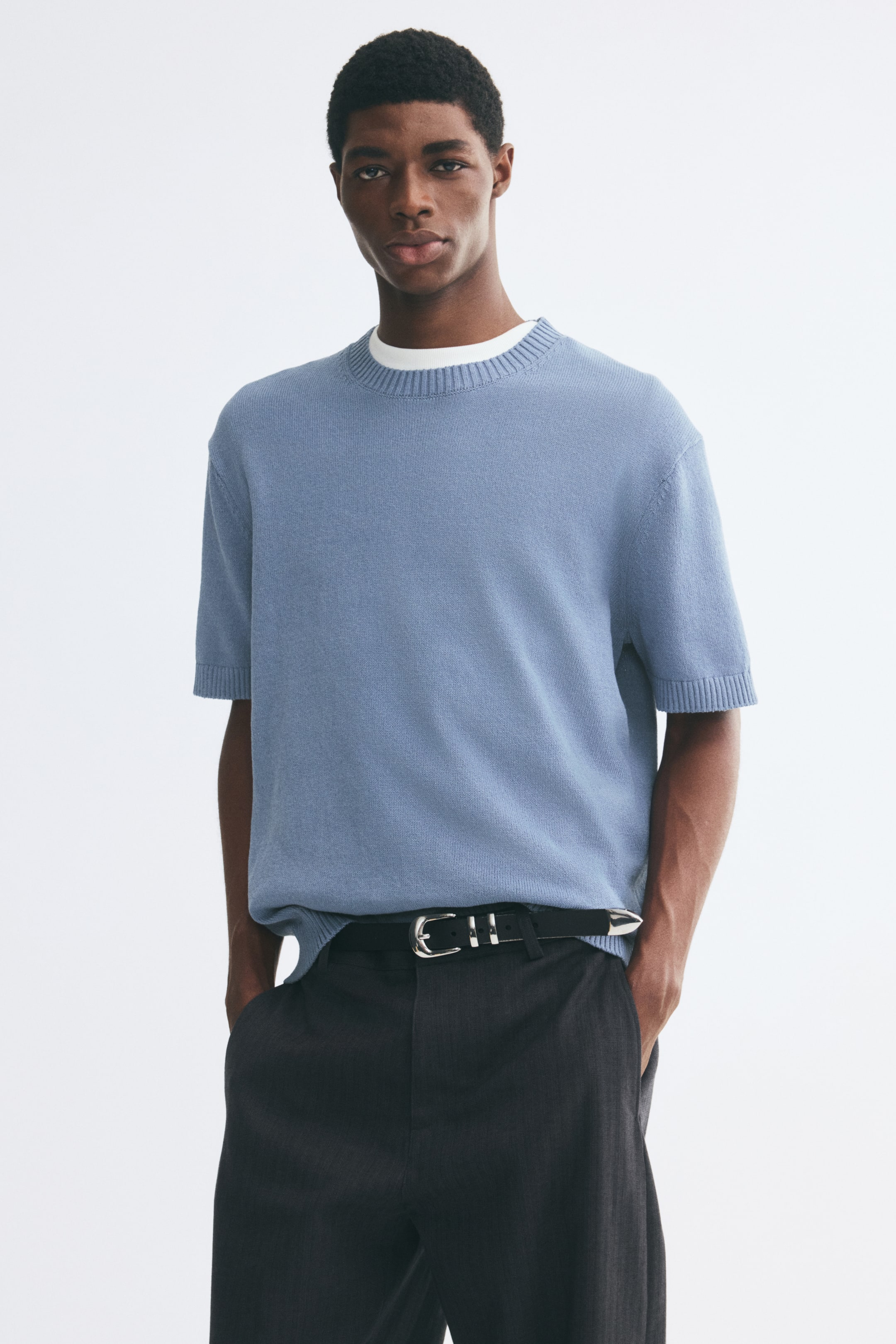 View larger image: The model is wearing a light blue, short-sleeve knit T-shirt with a ribbed crew neckline and ribbed cuffs. It is layered over a white crewneck undershirt and styled with dark trousers.