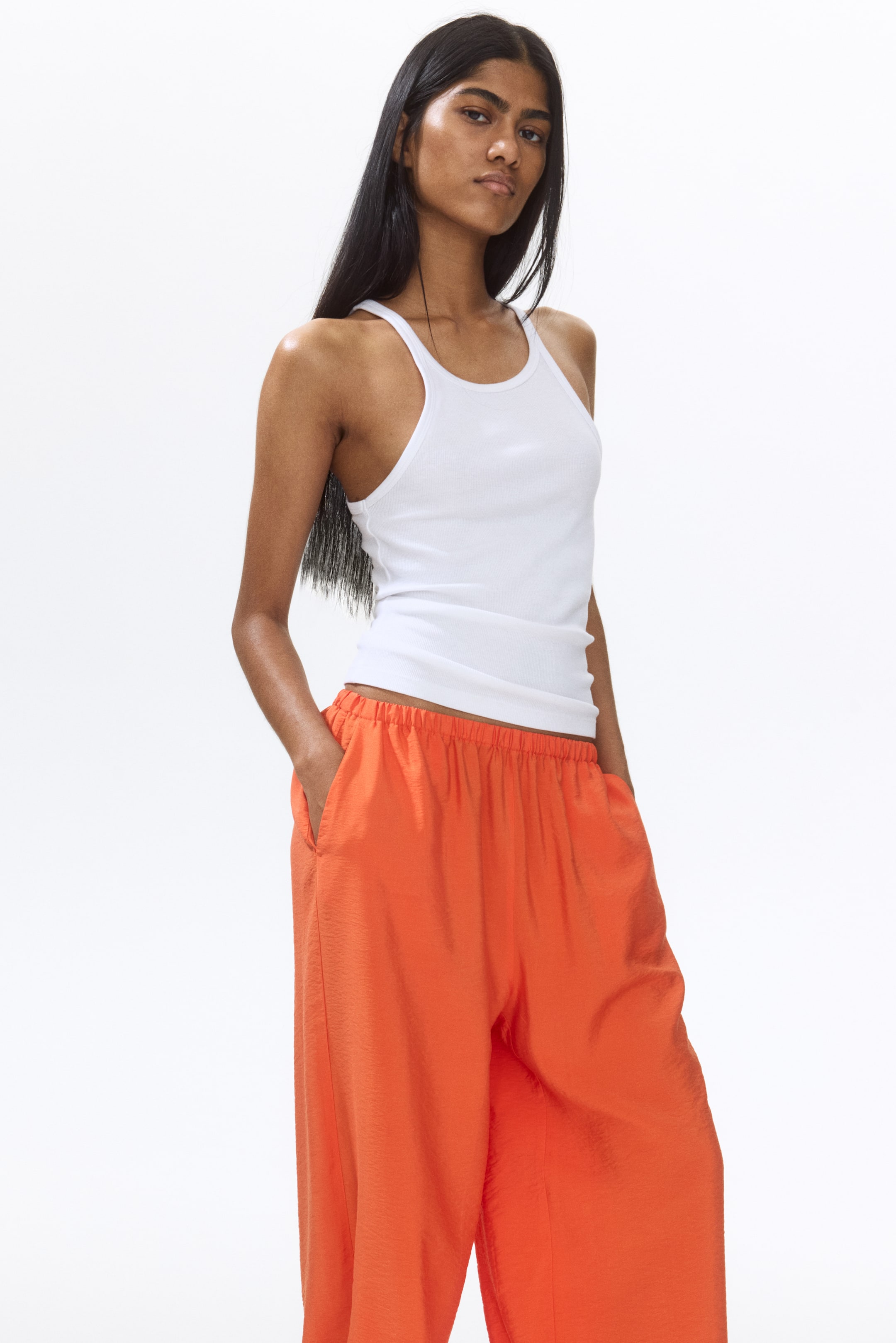 View larger image: The model is showcasing orange loose-fit trousers crafted from a viscose-blend weave, characterized by balloon legs. These trousers come with a concealed elasticated waistband and diagonal side pockets, with the model's hand casually placed in one of the pockets.