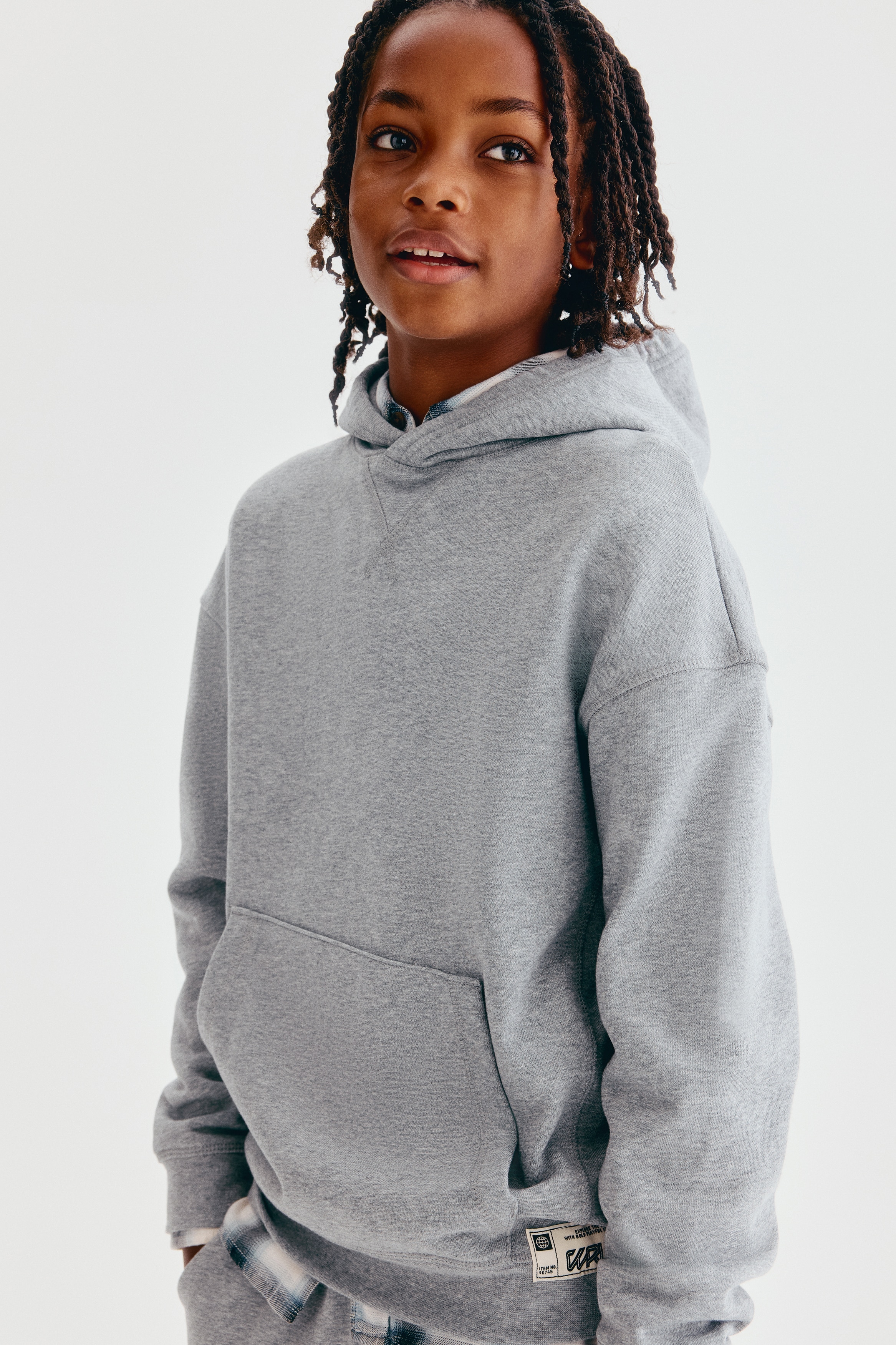 This loose-fit hoodie in heather grey is shown from the front, featuring a crossover hood, a V-stitch detail at the neckline, and dropped shoulders. It has a front kangaroo pocket, ribbed cuffs, and a ribbed hem with a small woven label at the bottom right.