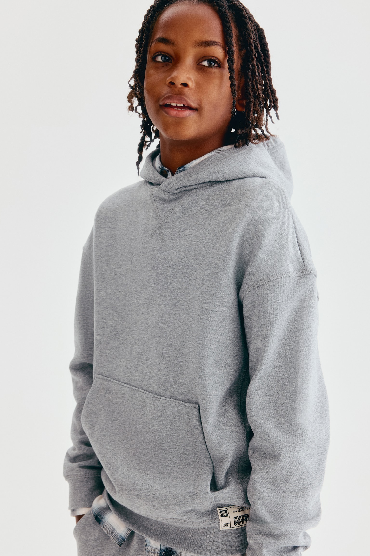 This loose-fit hoodie in heather grey is shown from the front, featuring a crossover hood, a V-stitch detail at the neckline, and dropped shoulders. It features a front kangaroo pocket, ribbed cuffs, and a ribbed hem with a small woven label on the bottom right. - 1