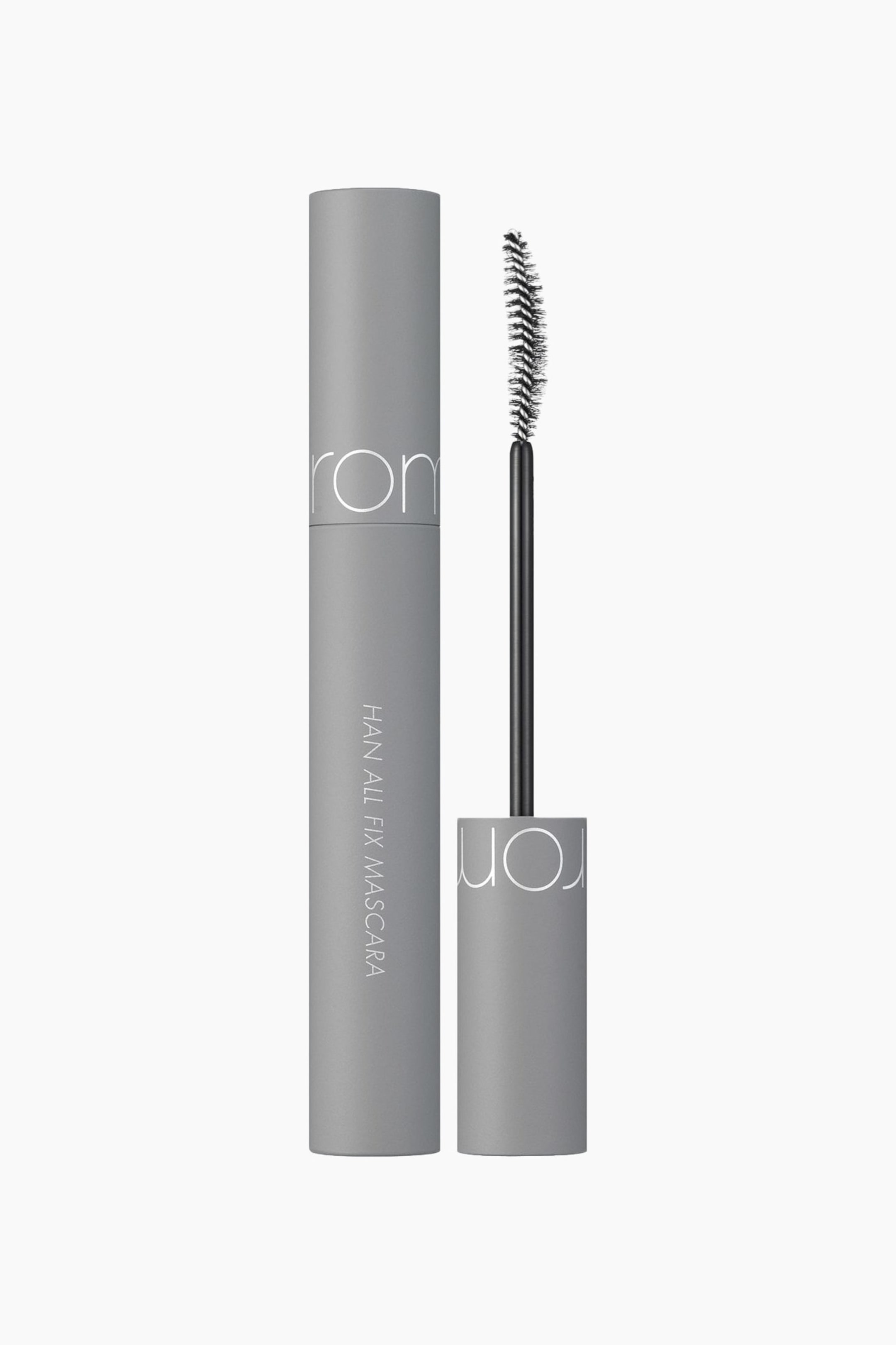 Vis større bilde: En grå mascara presenteres med det sylindriske hylsteret stående oppreist, med «rom&nd» horisontalt og «HAN ALL FIX MASCARA» vertikalt. Den forlengede svarte kosten, med et grått skaft med «rom&nd»-tekst, viser en buet svart børste.