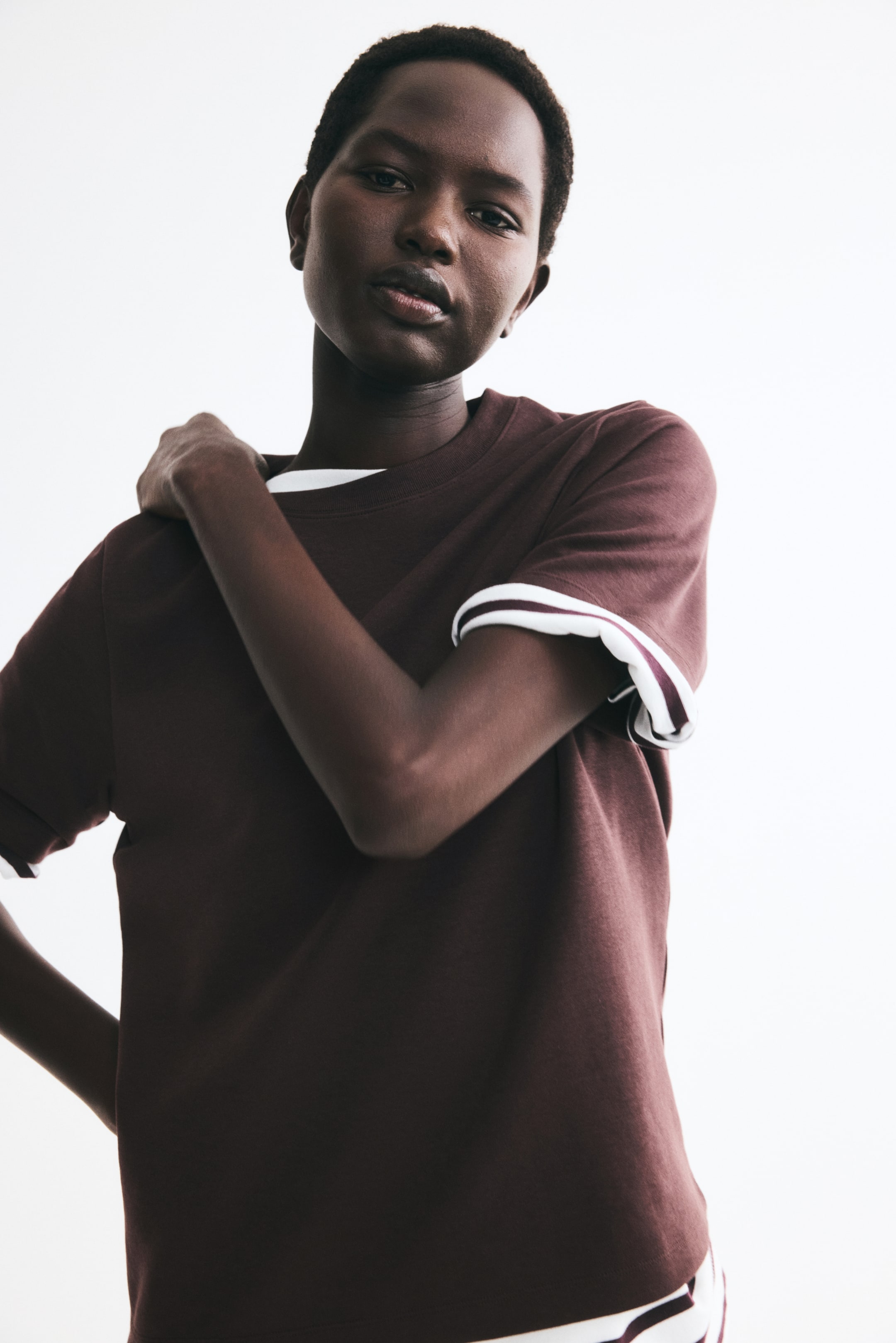 Büyük resmi görüntüle: Model, kahverengi kalın interlock jersey bir t-shirt giyiyor; yuvarlak, ribana detaylı yaka, yakadan görünen beyaz bir t-shirt üzerine katmanlı şekilde kombinlenmiş. Katlanmış kol manşetleri ve etek ucuyla öne çıkan parça, düz kesim eteği ve yapılandırılmış kalıbı vurgulamak için model bir elini karşı omzuna yaslıyor.