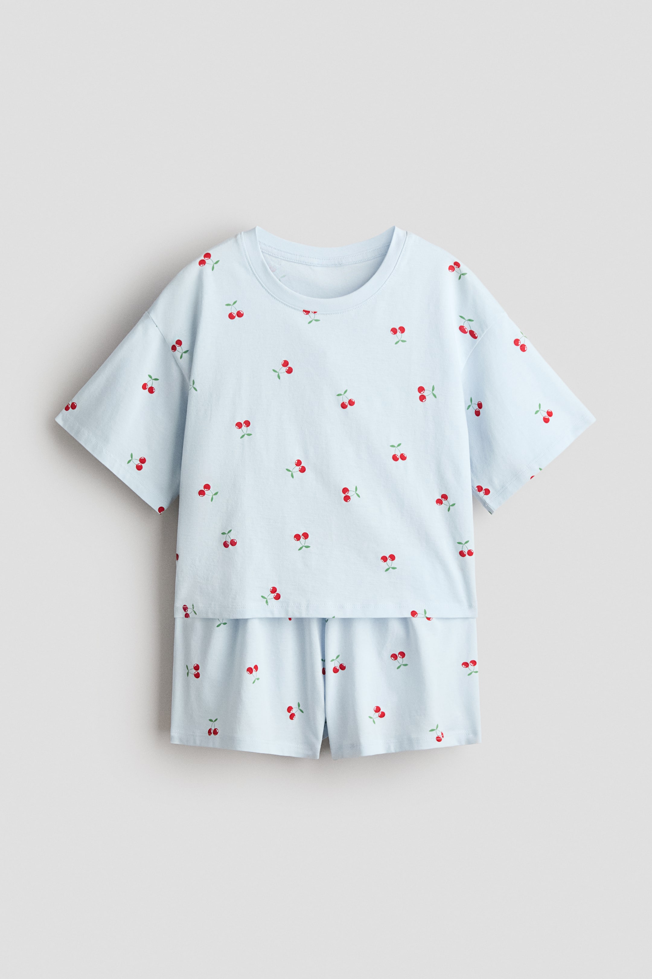 Agrandir l'image: Un ensemble pyjama en jersey de coton bleu clair présente un imprimé de cerises rouges éparpillées, vu de face. Le haut est doté d'un col rond, d'épaules tombantes et de manches courtes, tandis que le short est conçu avec une taille élastiquée.