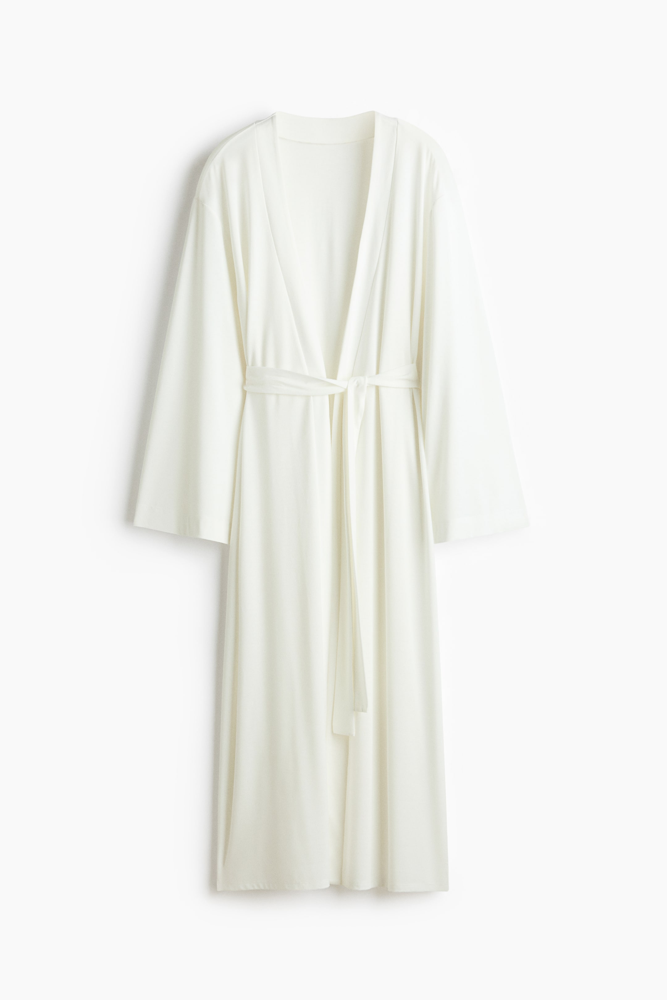 View larger image: The white robe showcases an open V-neckline and wide sleeves. A matching fabric belt ties at the waist, allowing the soft material to drape to approximately knee-length.