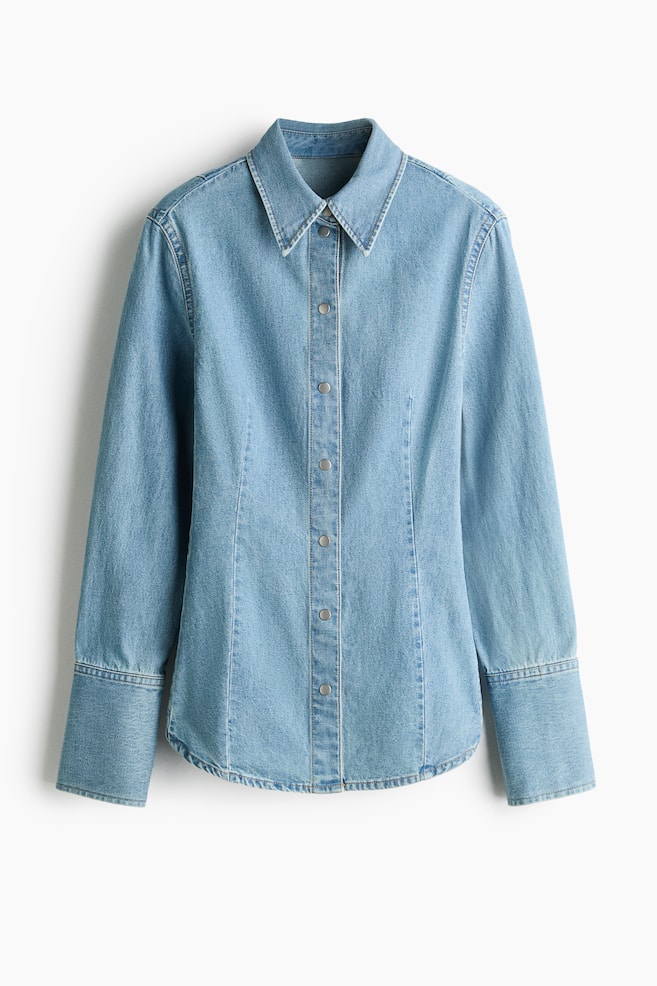 A light-wash blue denim shirt is shown from the front, featuring a structured point collar, a silver-tone snap-button front closure, and vertical contour seams. The long sleeves end in extra-wide cuffs, and the shirt tapers slightly at the waist before finishing with a curved hem. - 1