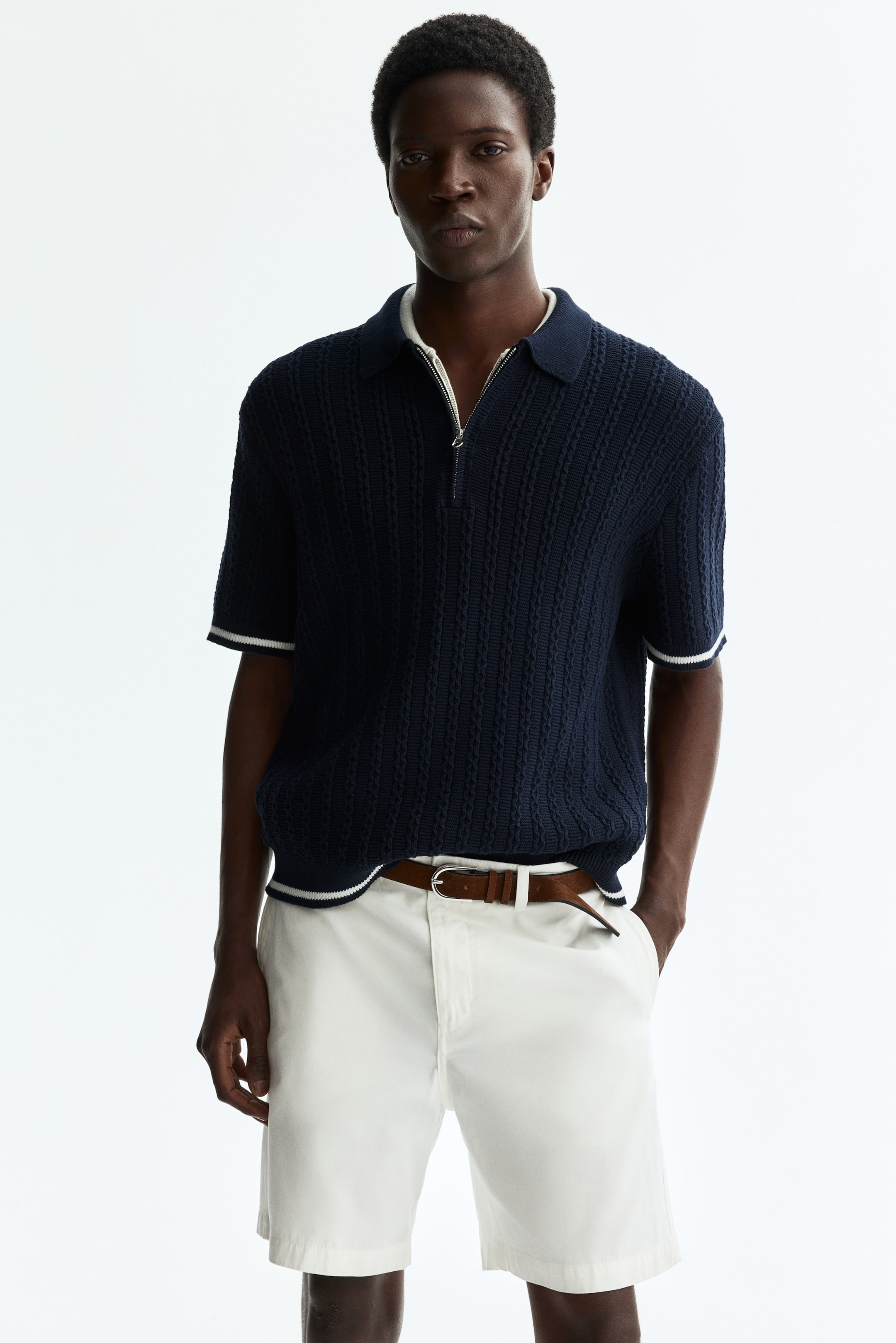 View larger image: A navy blue knitted polo shirt showcases a textured knit pattern, a polo collar, and a half-zip closure with a white undershirt peeking through beneath. It features short sleeves with ribbed cuffs and a ribbed hem, both highlighted with white stripes.