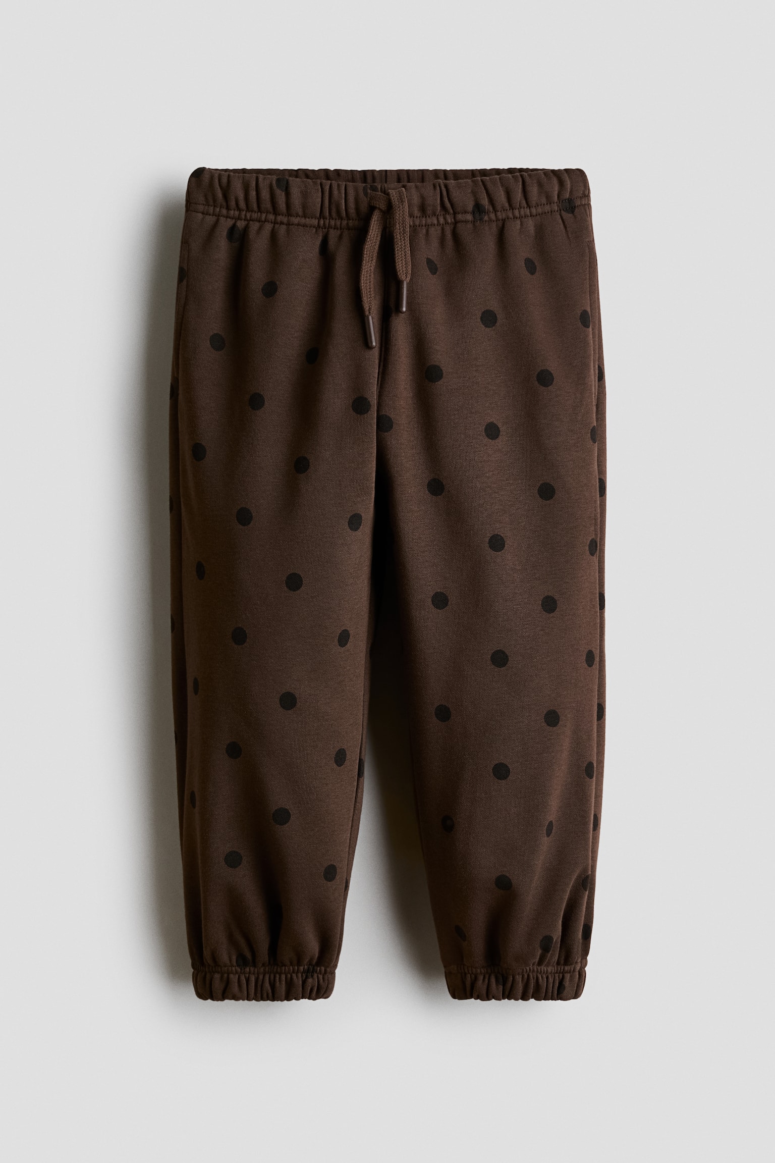 Showing a front view, these dark brown joggers feature an all-over black polka-dot pattern, an elasticated waistband with a matching drawstring, and gathered elastic cuffs.