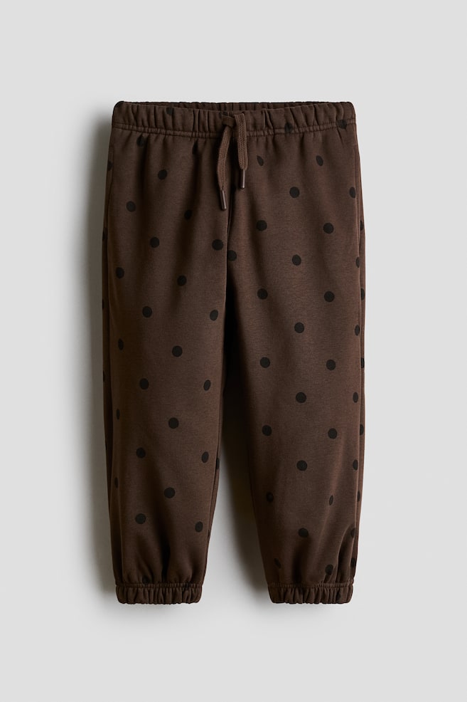 Showing a front view, these dark brown joggers feature an all-over black polka-dot pattern, an elasticated waistband with a matching drawstring, and gathered elastic cuffs. - 1