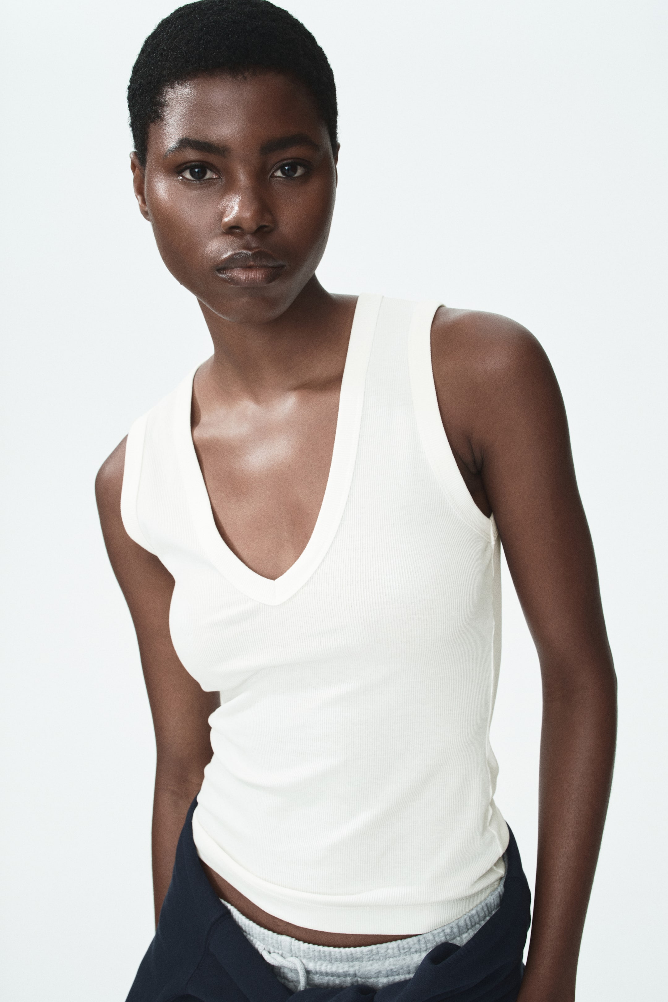 View larger image: The model is sporting an off-white, ribbed jersey vest top that features a fitted silhouette, a deep V-neckline, and a narrow trim around the neckline and armholes. The top is styled with trousers that sit low on the hips.