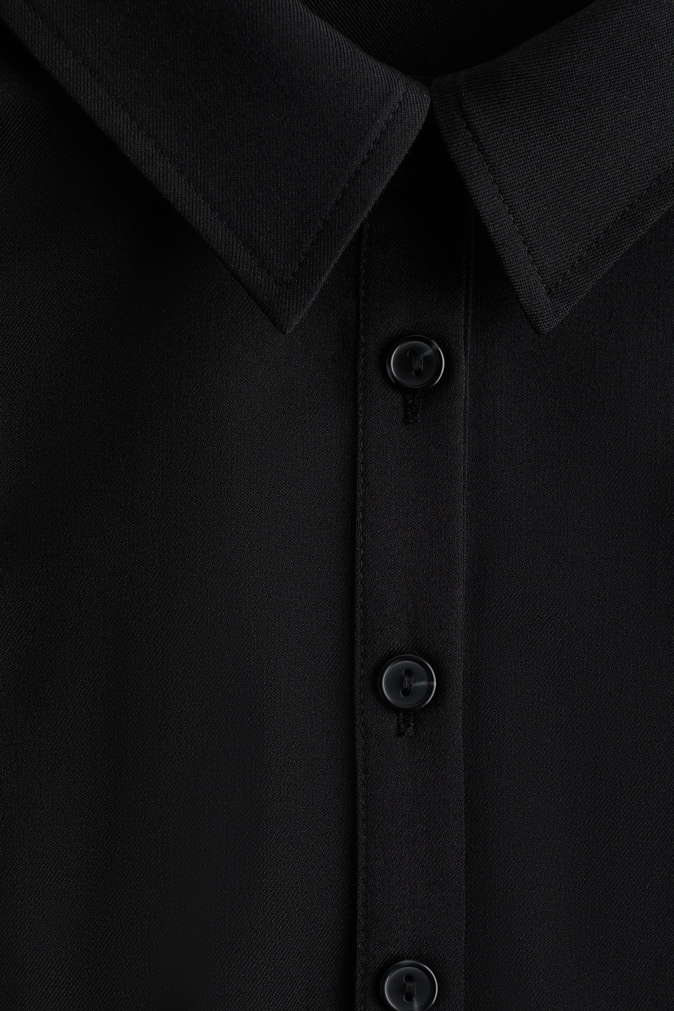 拡大画像を表示: 黒の織りシャツがフロントから紹介されています。このシャツは、クラシックなポイントカラーが特徴で、ステッチが見えるデザインになっており、三つの黒の丸い四つ穴ボタンが付いたボタン付きプラケットがあります。