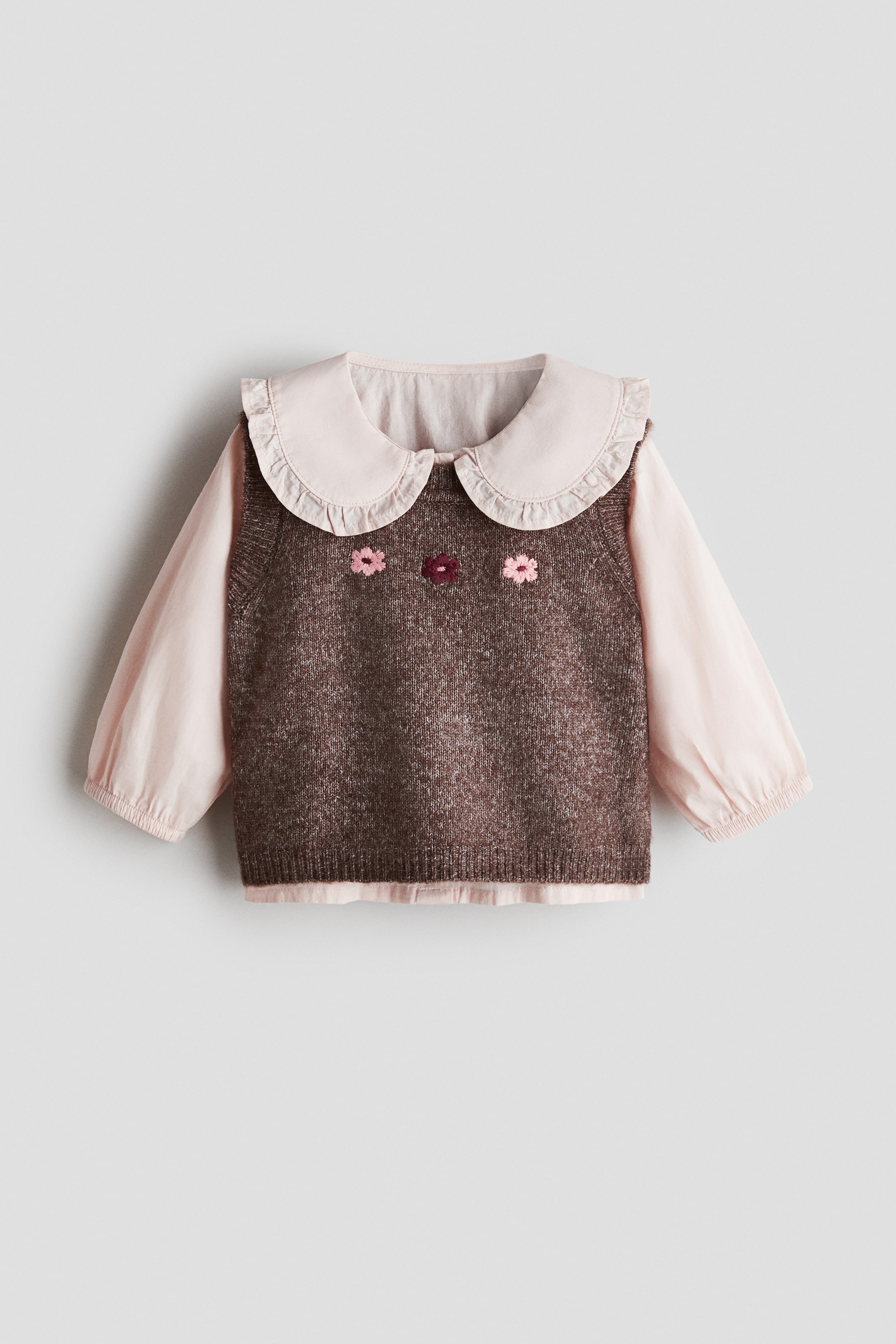 Ver imagem maior: Um colete de malha castanho, com três flores bordadas na frente, está sobreposto a uma camisola de manga comprida cor-de-rosa claro, apresentada de frente. A camisola exibe uma grande gola Peter Pan com folhos e mangas compridas com punhos elásticos.
