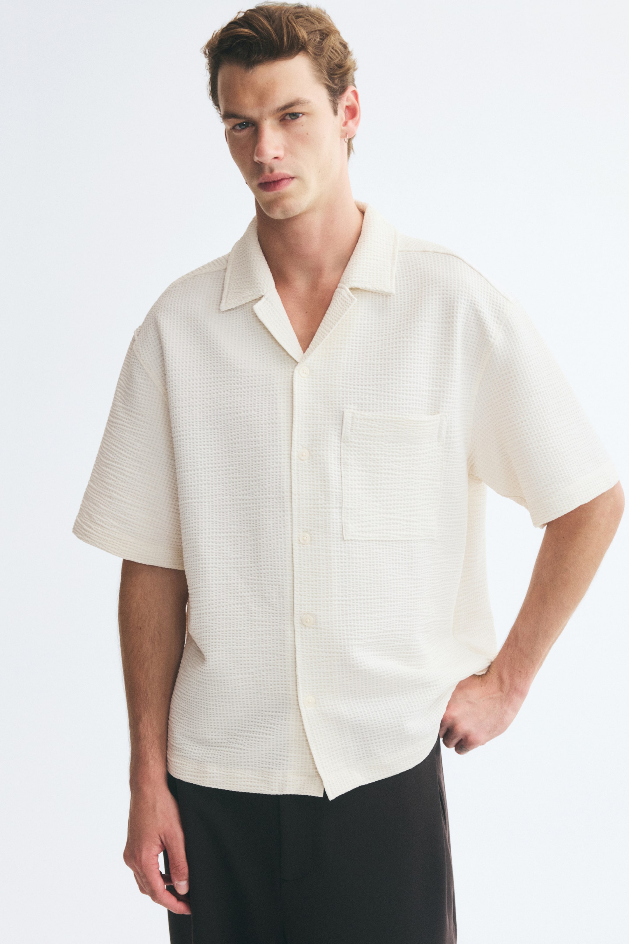 Voir une image plus grande: Une chemise à manches courtes en mélange de coton à texture gaufrée blanc cassé est portée par le mannequin. La chemise présente une coupe décontractée, un col camp, une seule poche poitrine et un ourlet droit, et elle est agencée avec un pantalon foncé.
