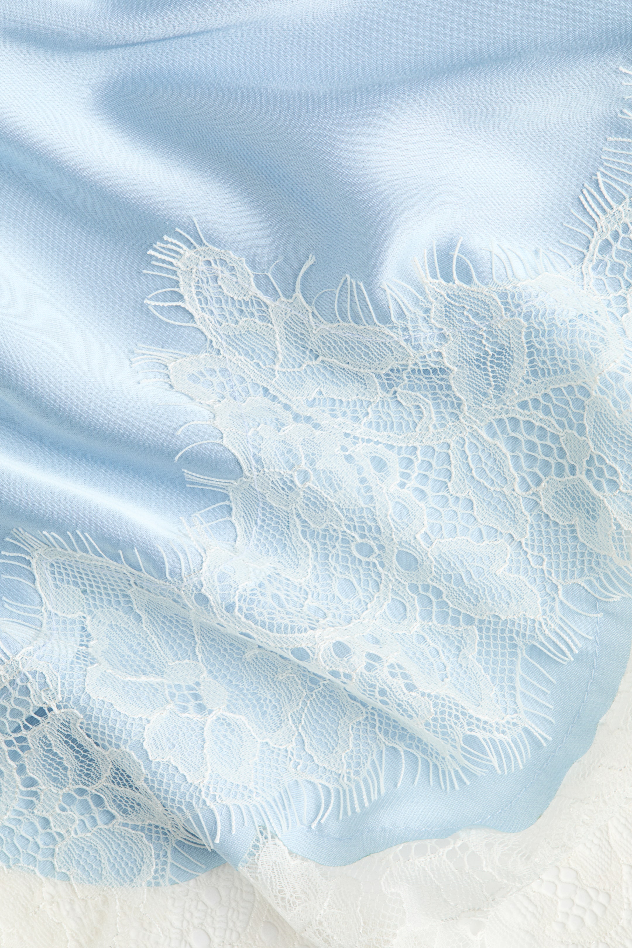 Visualizza l'immagine ingrandita: Un ampio foulard triangolare in raso azzurro chiaro, che cade morbidamente, mostra il suo davanti, caratterizzato da un bordo in pizzo bianco lungo due lati.