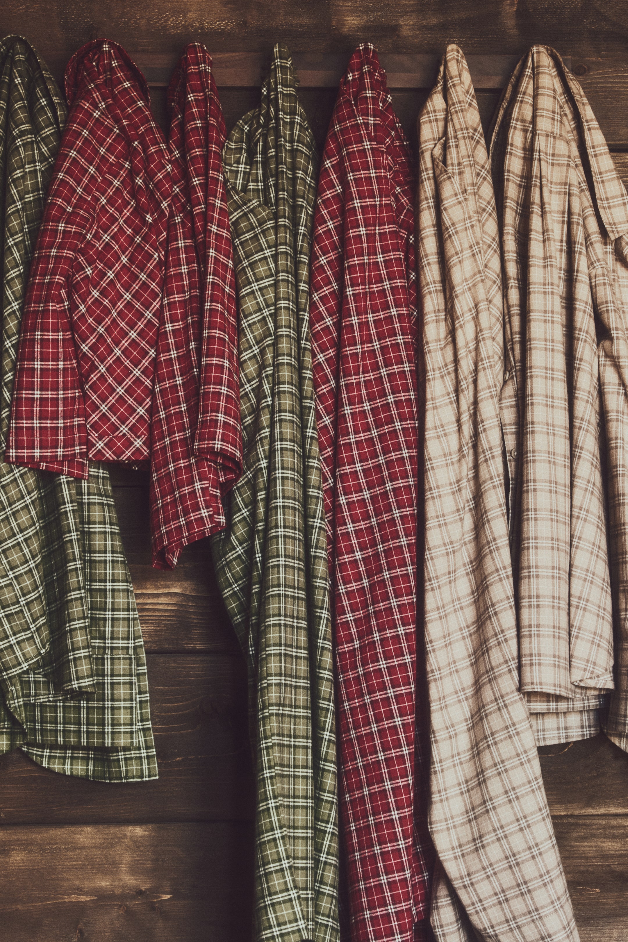 View larger image: Several long-sleeved plaid shirts in shades of red, green, and beige are hanging vertically, showcasing their checkered patterns and woven textures. The shirts are draped closely together, displaying various folds, collars, and button details.