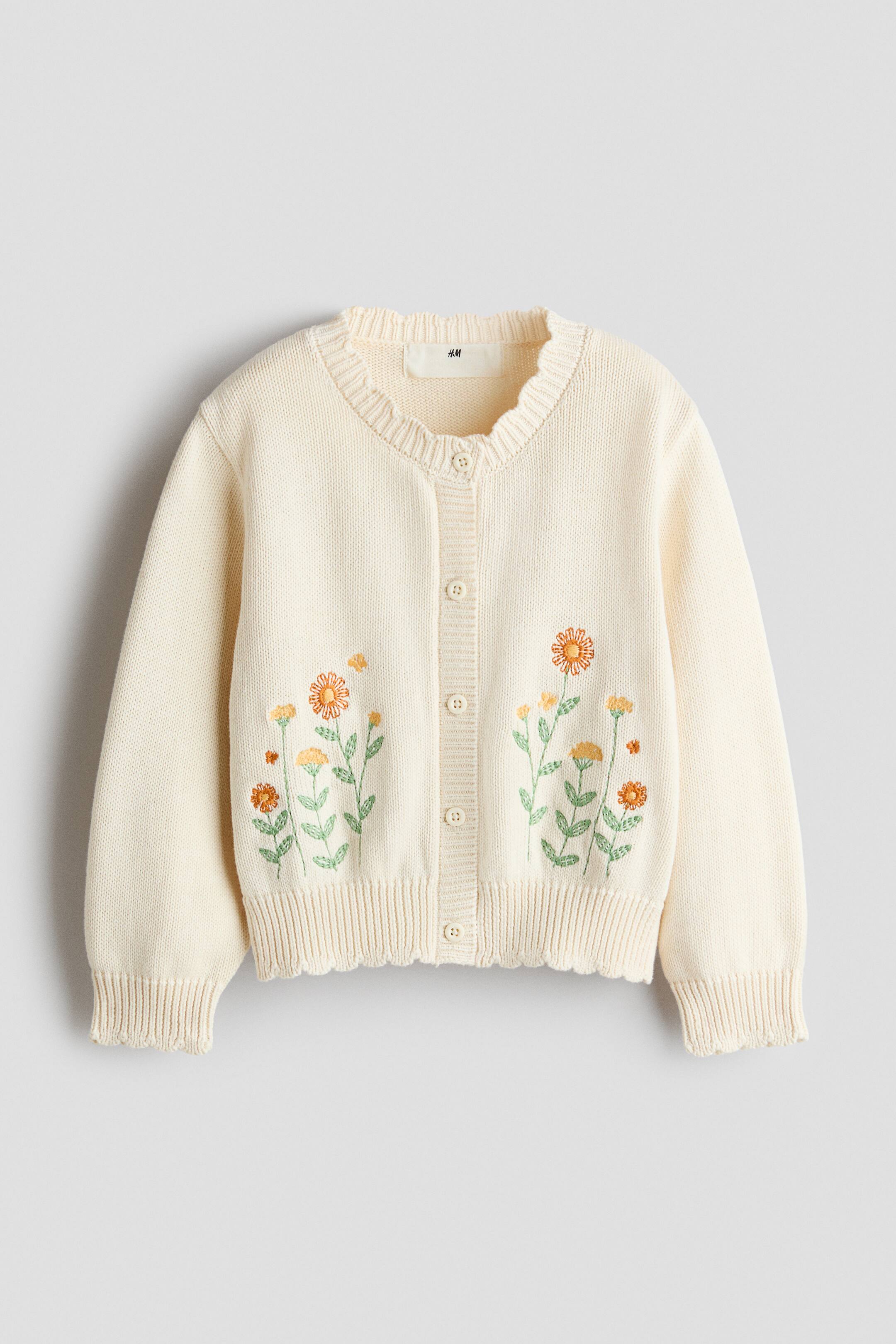 View larger image: A cream-colored cotton knit cardigan is shown from the front, featuring embroidered orange and yellow floral motifs on the lower front. It has a round neckline, a button-down front, dropped shoulders, and ribbed trims with scalloped edges at the neckline, cuffs, and hem.