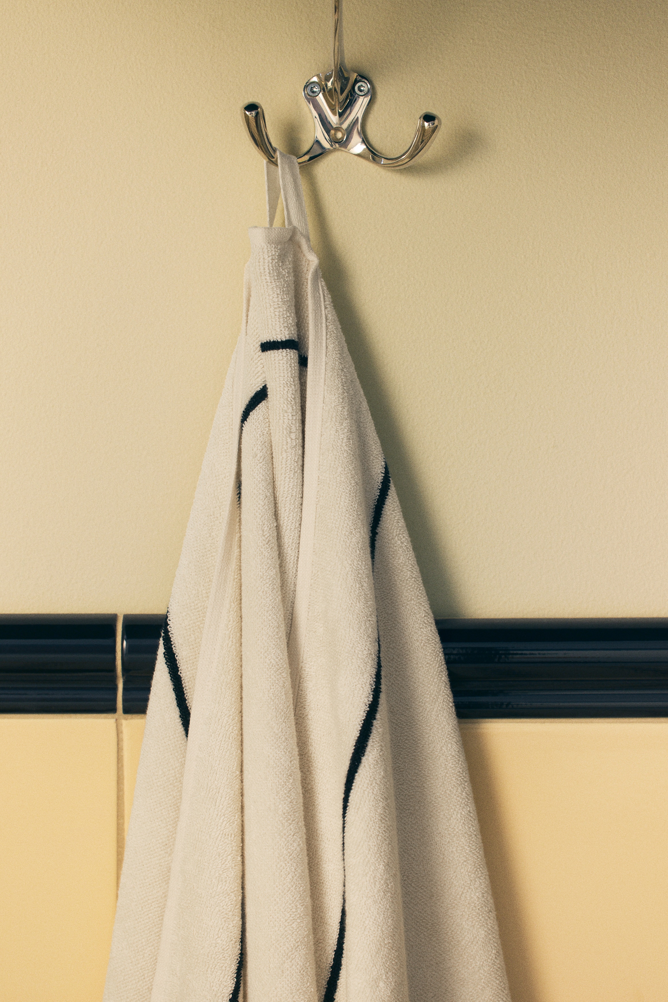 An off-white towel is hanging from a black hook, presenting its folded front surface. It is featuring a pattern of thin black lines forming an abstract grid design.