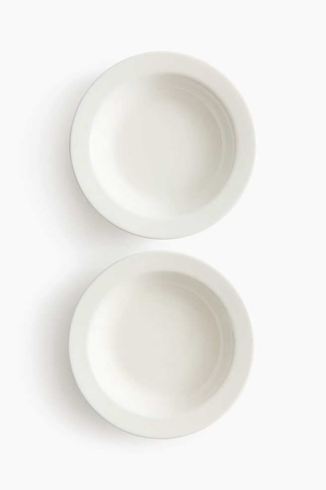 Deux bols ronds de couleur blanc cassé sont orientés vers le haut, l'un placé au-dessus de l'autre. Chaque bol présente un large rebord plat et un centre peu profond et creusé. - 1