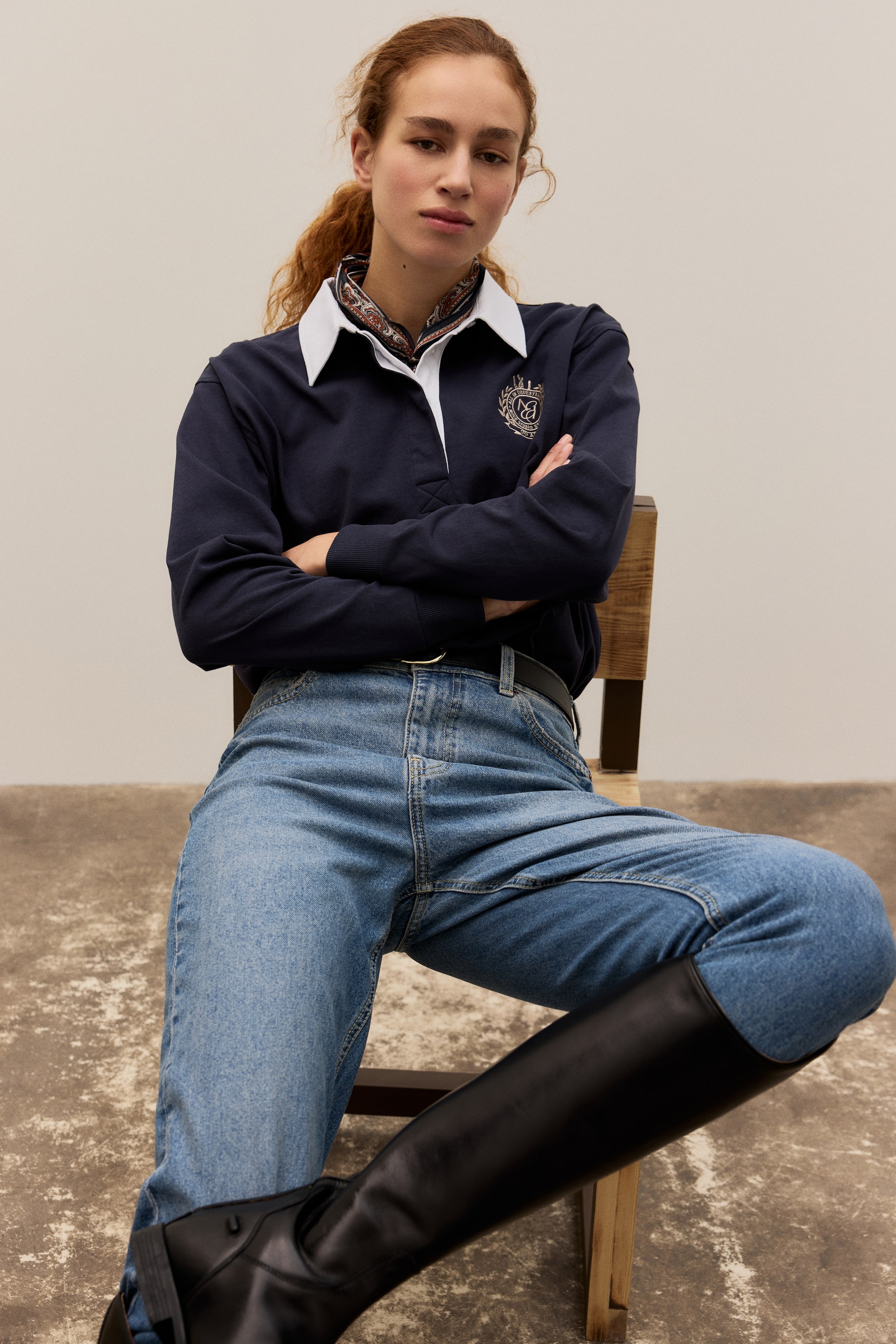 Visa större bild: En marinblå långärmad rugbytröja med vit krage, dold vit knappslå och ett guldbroderat emblem på vänster bröst, med en mönstrad scarf under kragen. Den kombineras med ljusblå jeans, säkrade med ett svart bälte, och höga svarta läderridstövlar.
