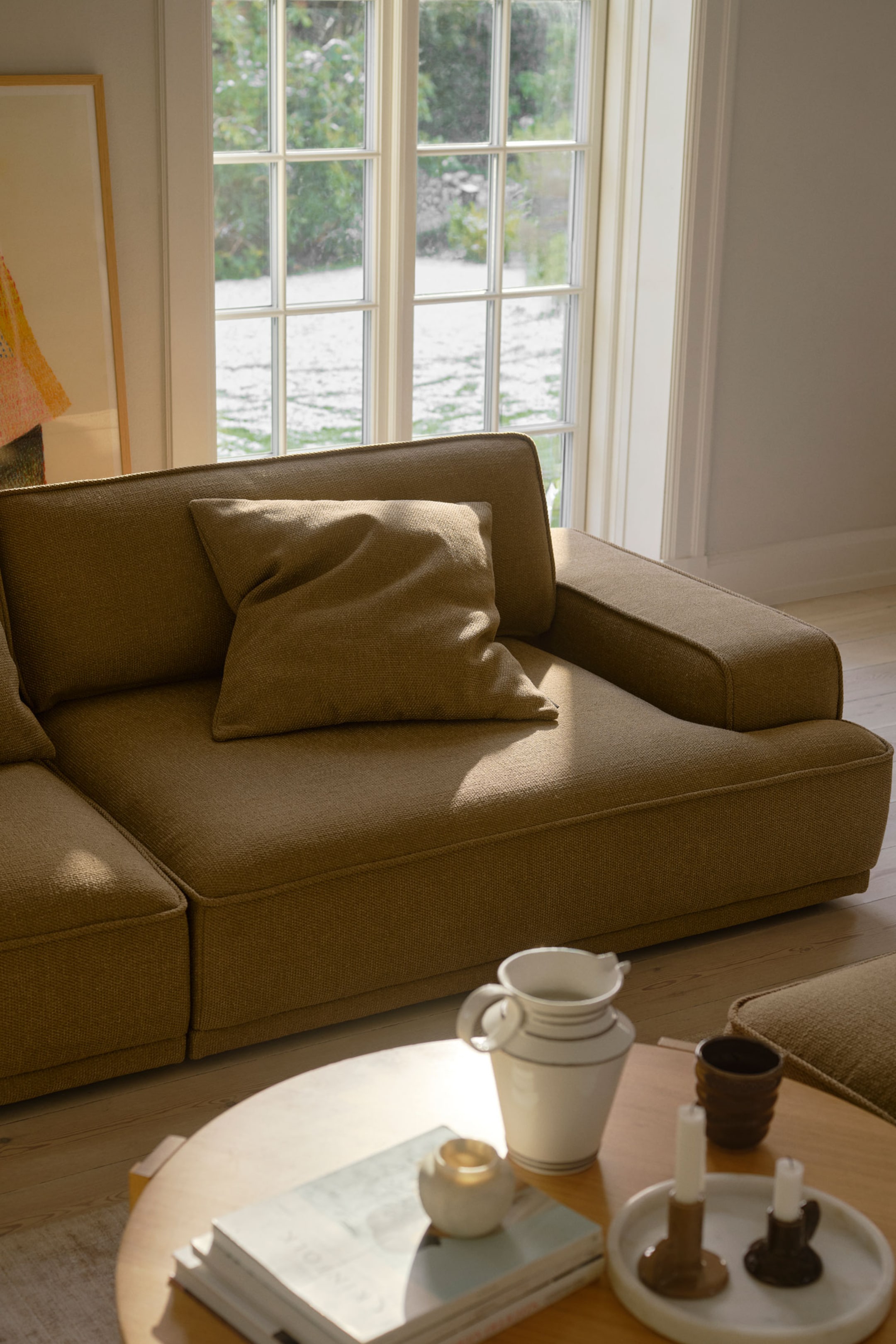 Größeres Bild ansehen: Ein niedriges, modulares Sofa, bezogen mit einem strukturierten, ockerfarbenen Stoff, ist nach rechts ausgerichtet und betont die tiefe Sitzfläche, die niedrige Rückenlehne und die breiten, kantigen Armlehnen. Ein passendes quadratisches Zierkissen lehnt auf dem Sitzpolster an der Rückenlehne.