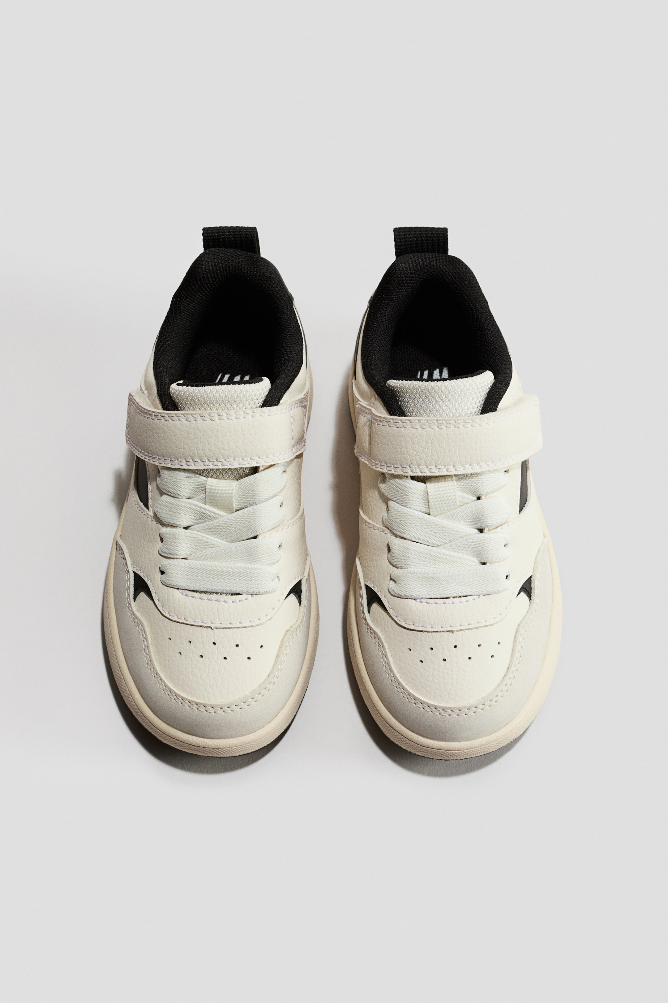 Agrandir l'image: Baskets. Une paire de baskets de couleur crème est présentée de face, avec des lacets plats blanc cassé et une fermeture à velcro assortie. Elles présentent une doublure intérieure noire, une languette de tirage noire au talon et des détails perforés sur la boîte à orteils.
