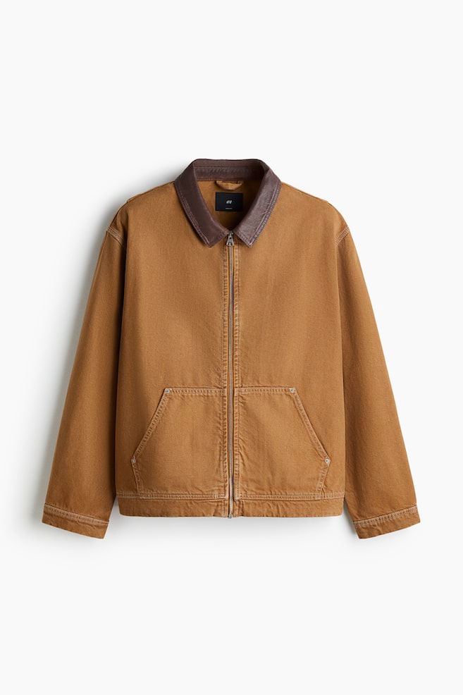 Eine kamelbraune Jacke aus einem robusten Stoff wird von vorne präsentiert, mit einem dunkelbraunen kontrastierenden Kragen und einem durchgehenden Metallreißverschluss. Zwei große aufgesetzte Taschen sind an der unteren Vorderseite sichtbar, jede mit schrägen Öffnungen und verstärkten Ecken. - 1