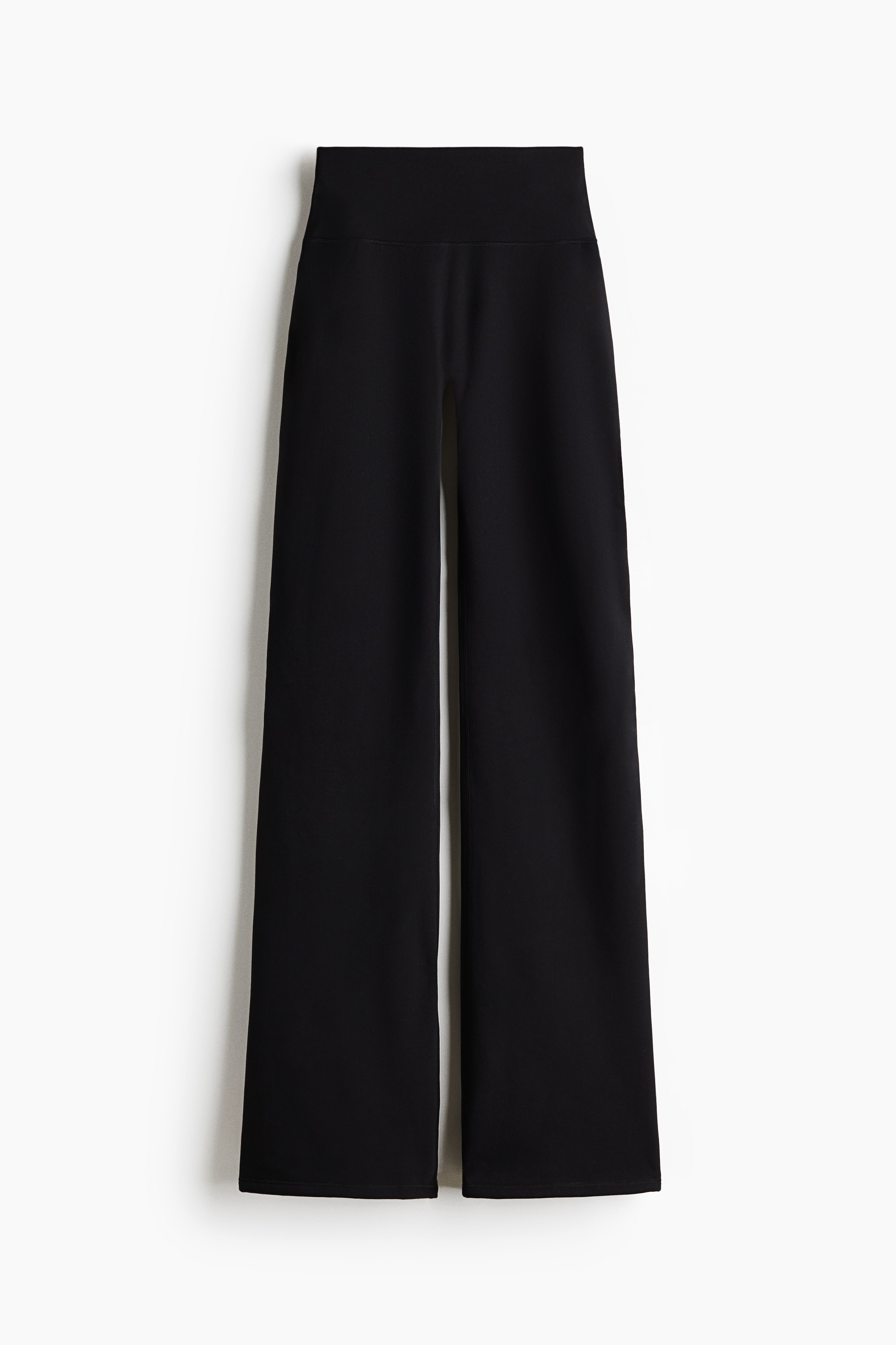 The model is wearing a black, oversized knitted turtleneck sweater paired with matching flared knitted trousers. The sweater features a high neck and long sleeves, while the trousers are styled with black slip-on shoes with flat soles.